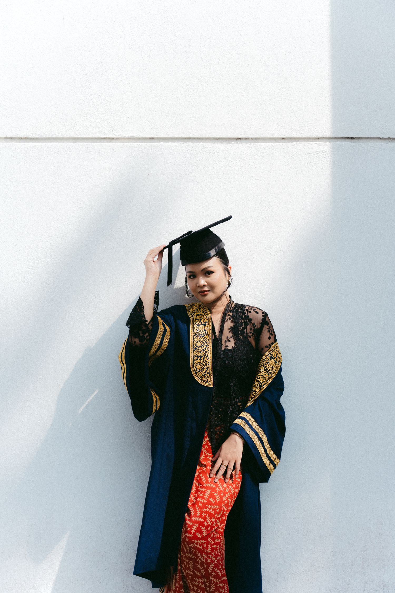 Graduate in black academic regalia with gold trim and red dress beneath poses against white wall during commencement.