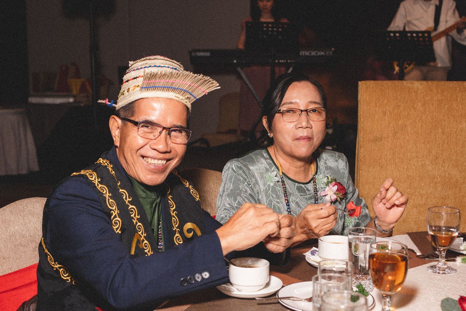 Two people having drinks at a restaurant table, with one wearing traditional embroidered clothing and glasses.