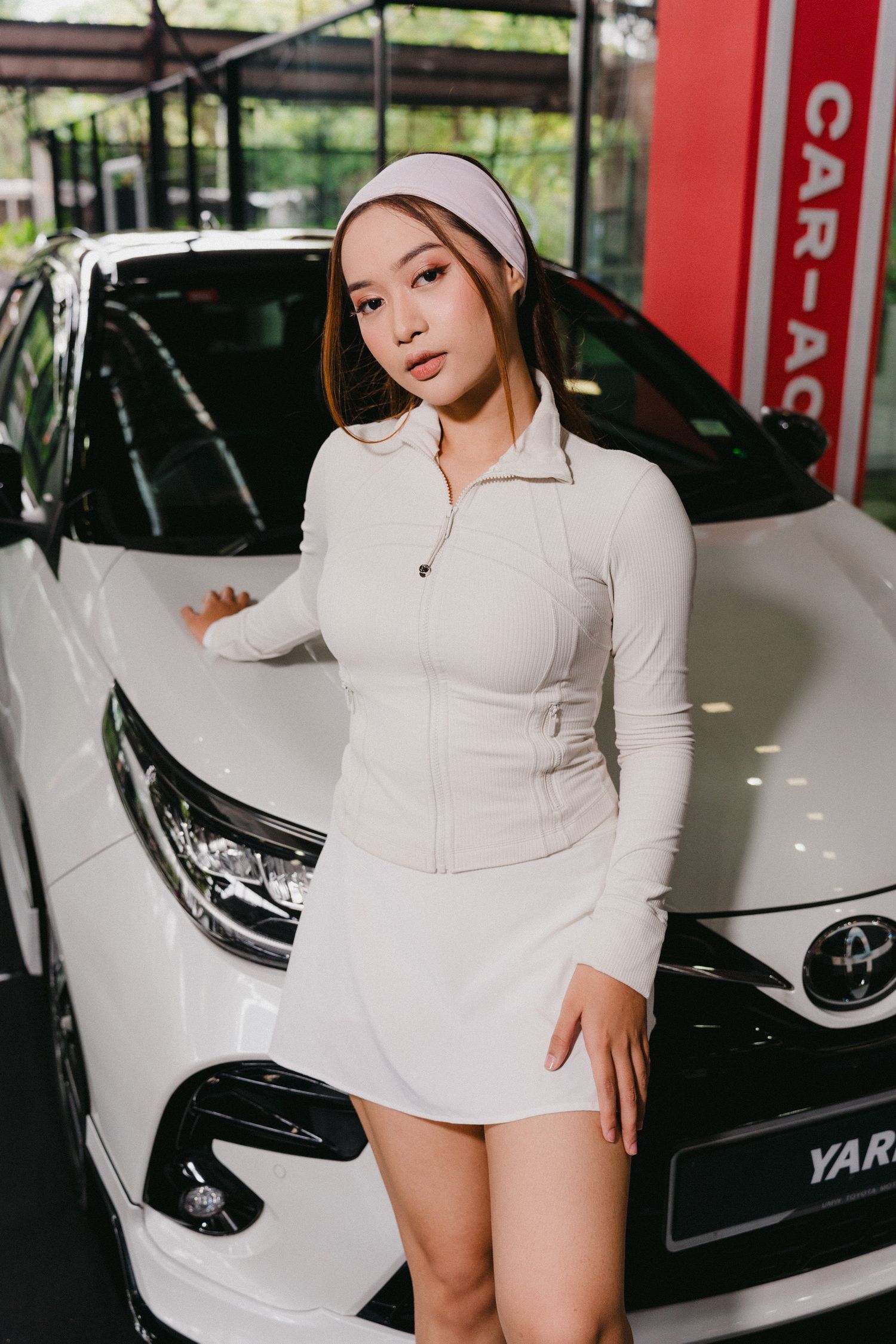 Woman in white outfit poses next to sleek white car in automotive showroom setting.