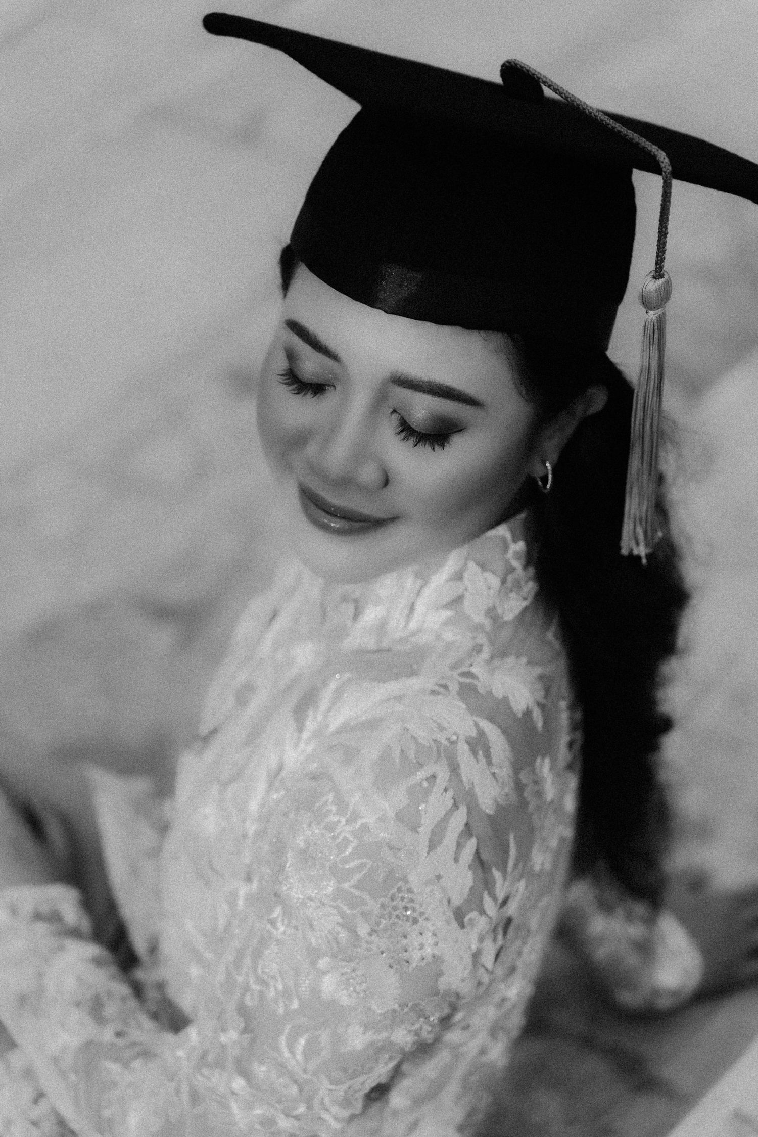A graduate in a cap and white lace dress smiles softly in a black and white portrait series.