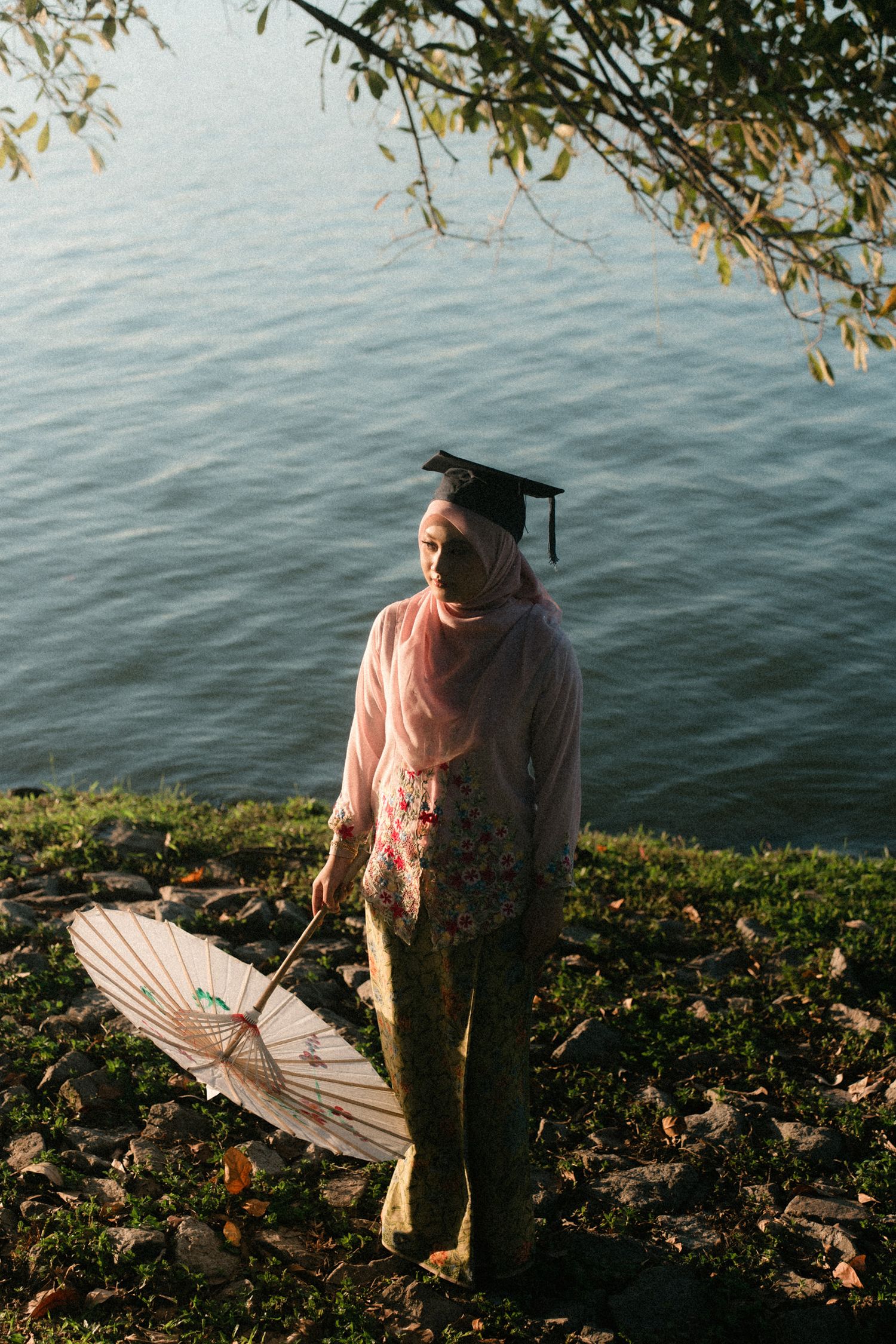 Graduate in a pink sweater and graduation cap stands by a lake holding a parasol on an autumn day.