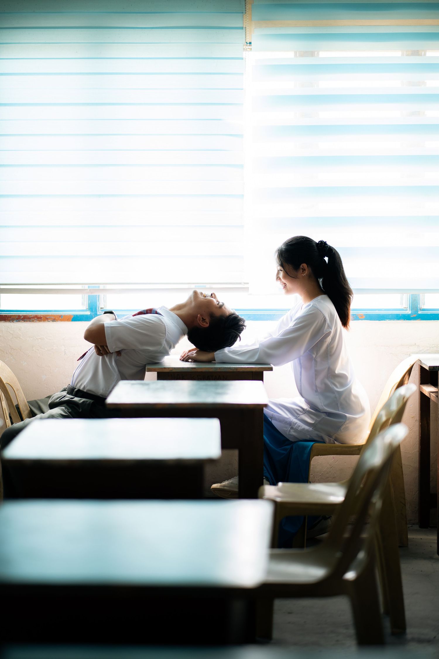 Students sitting at desks in a classroom with bright window light and venetian blinds in the background.