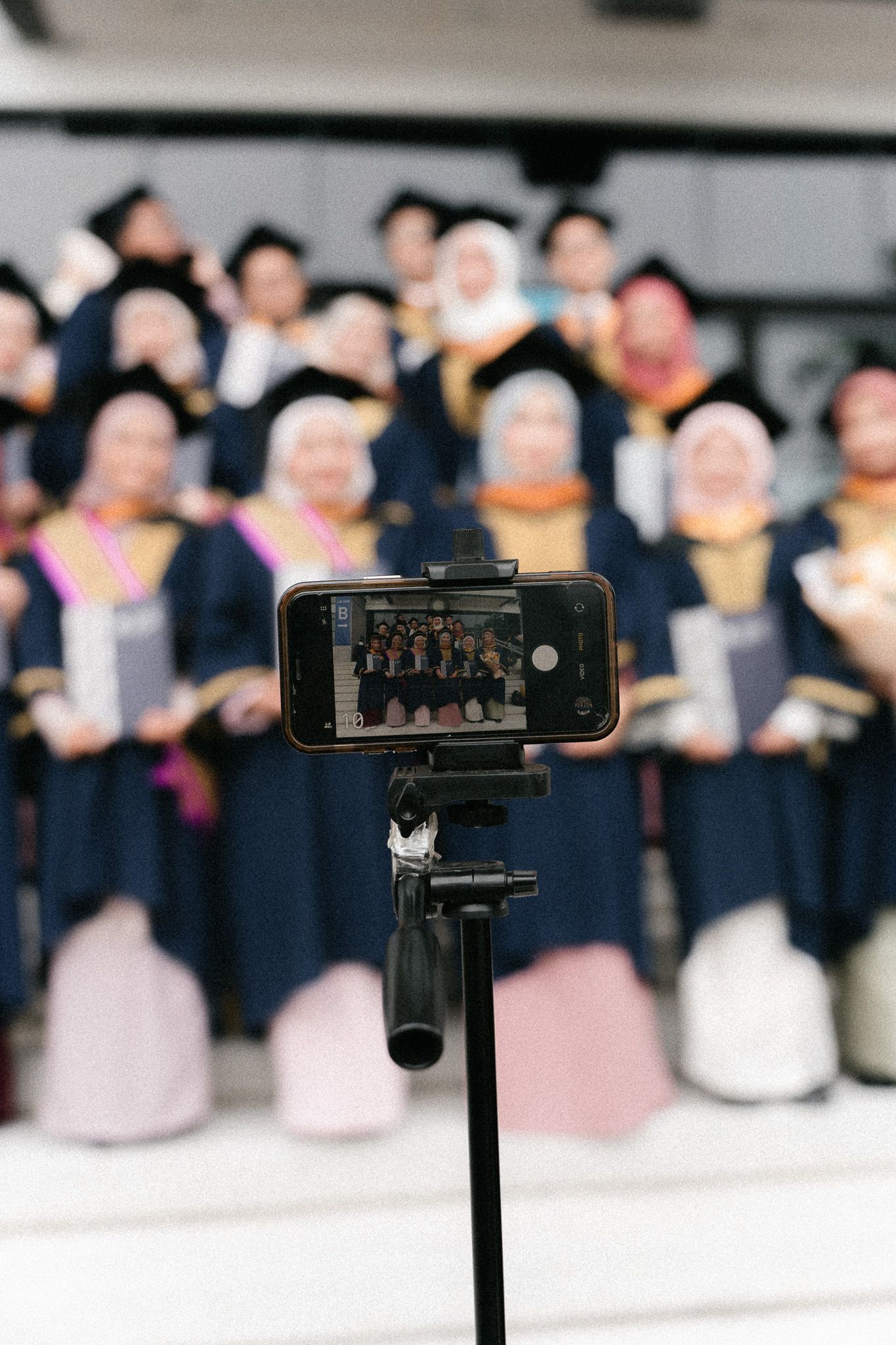Smartphone on tripod capturing group photo of graduates in blue robes and hijabs at graduation ceremony.