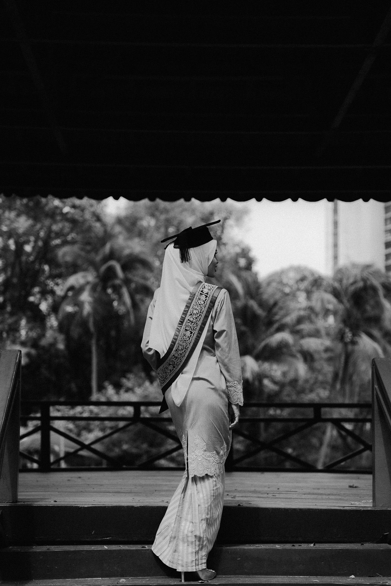 Black and white photo of a hijab-wearing graduate in traditional dress and mortarboard cap on outdoor steps.