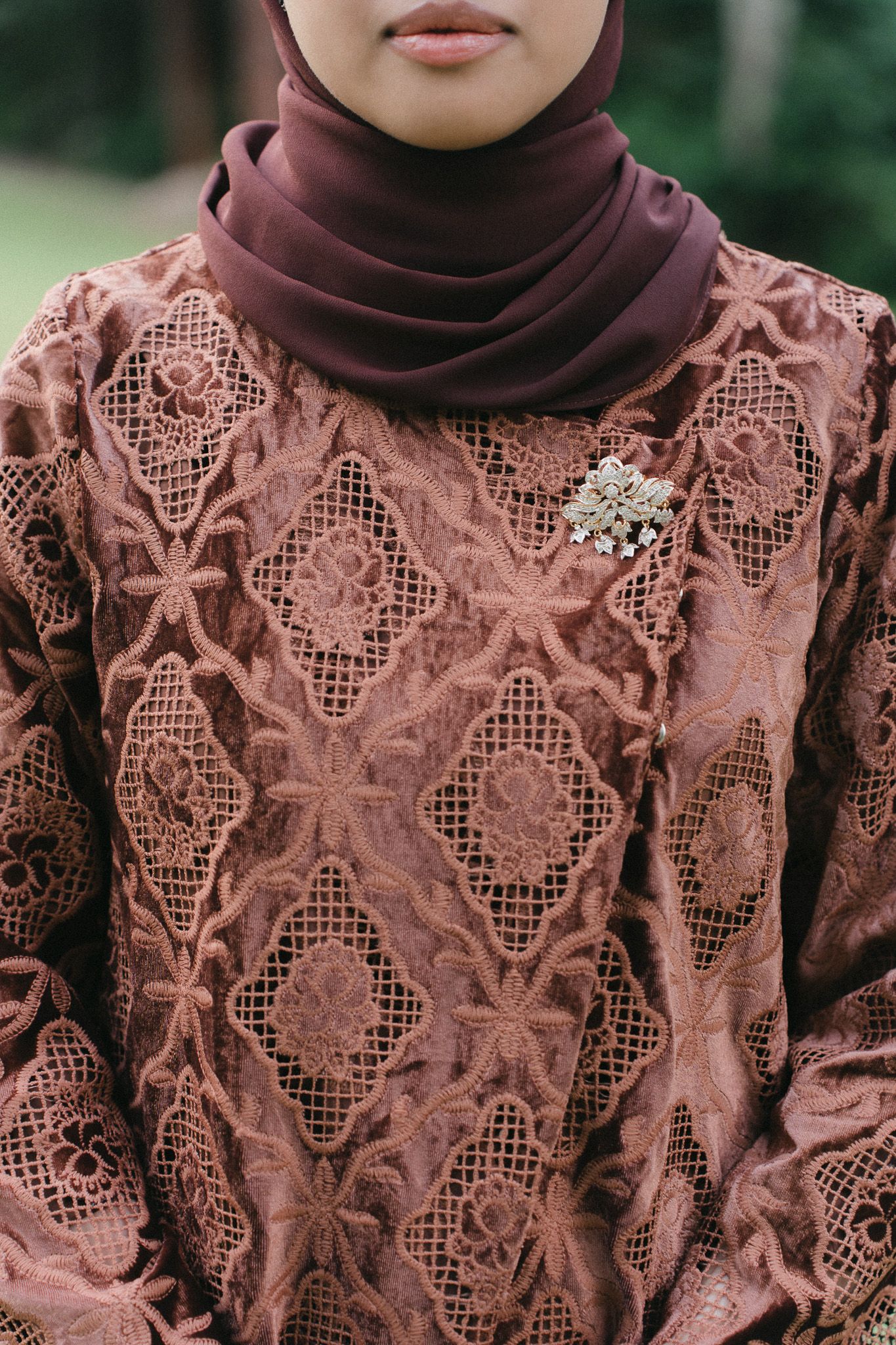Woman in dusty rose lace kebaya and maroon hijab with crystal brooch, elegant traditional Muslim fashion.