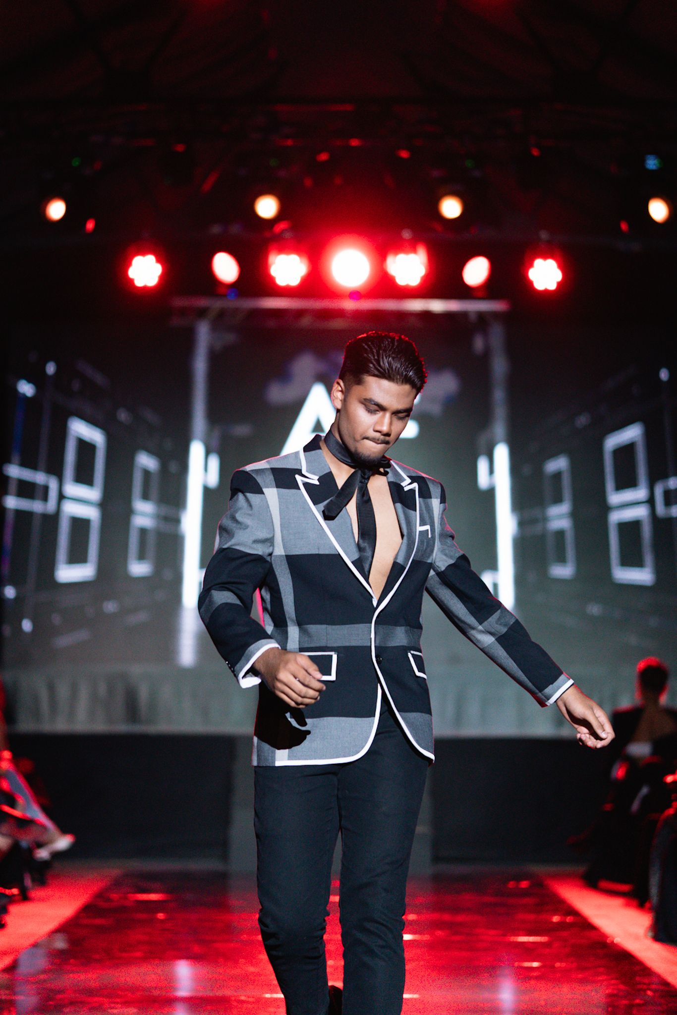 Model wearing structured grey and black blazer performs on red-lit runway.