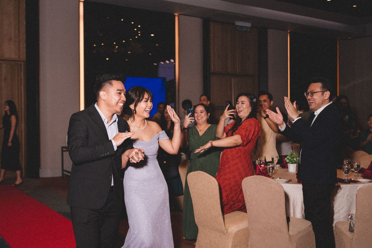 People dancing and celebrating at an elegant indoor wedding reception with decorative lighting and beige chair covers.