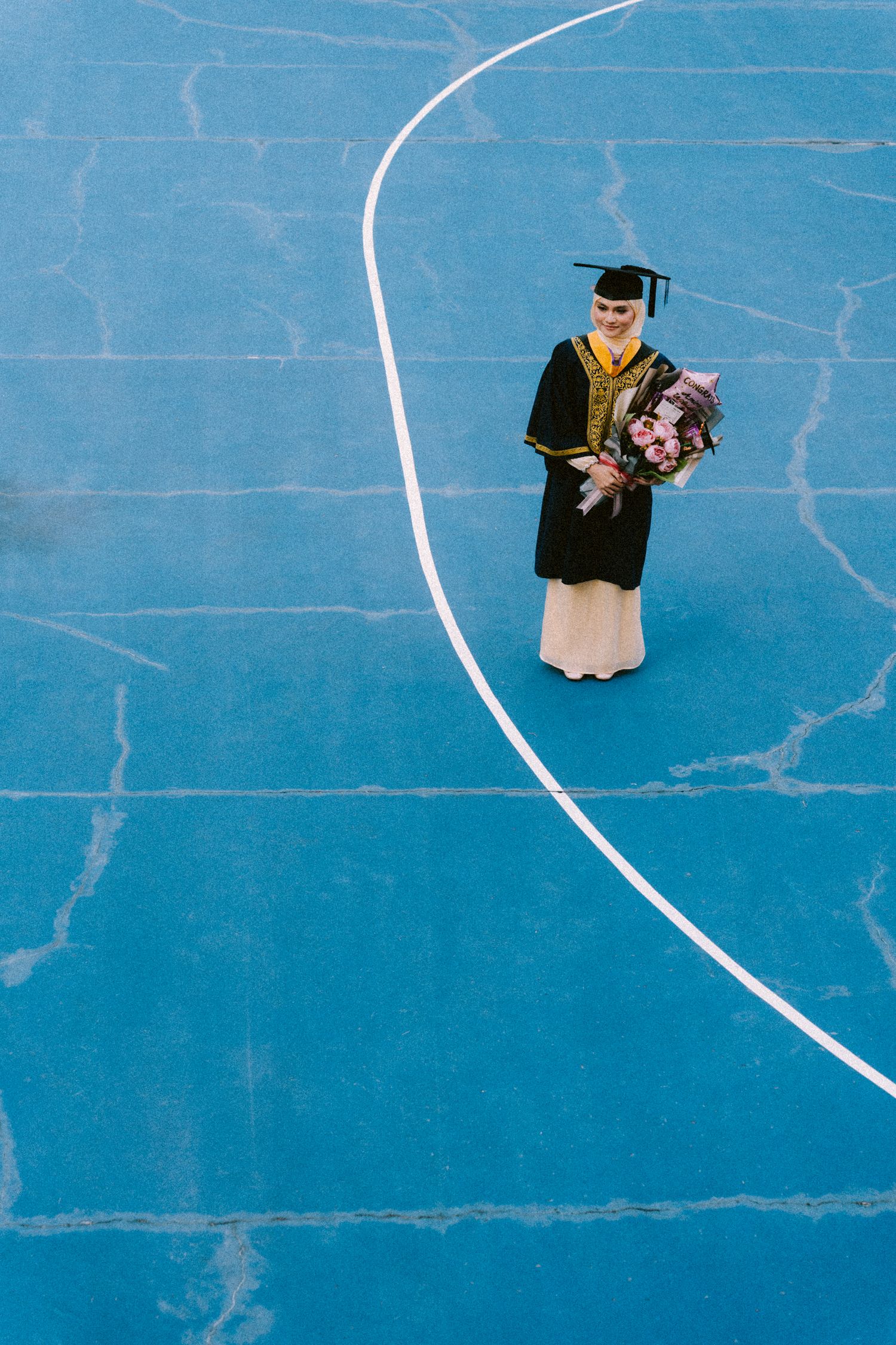 Graduate in black academic regalia holding flowers stands on blue court with white curved line.