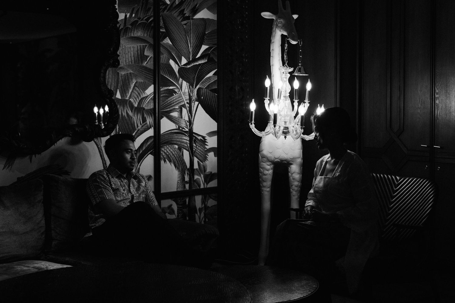 A moody black and white photo in a dark lounge setting with silhouettes and a glowing chandelier.