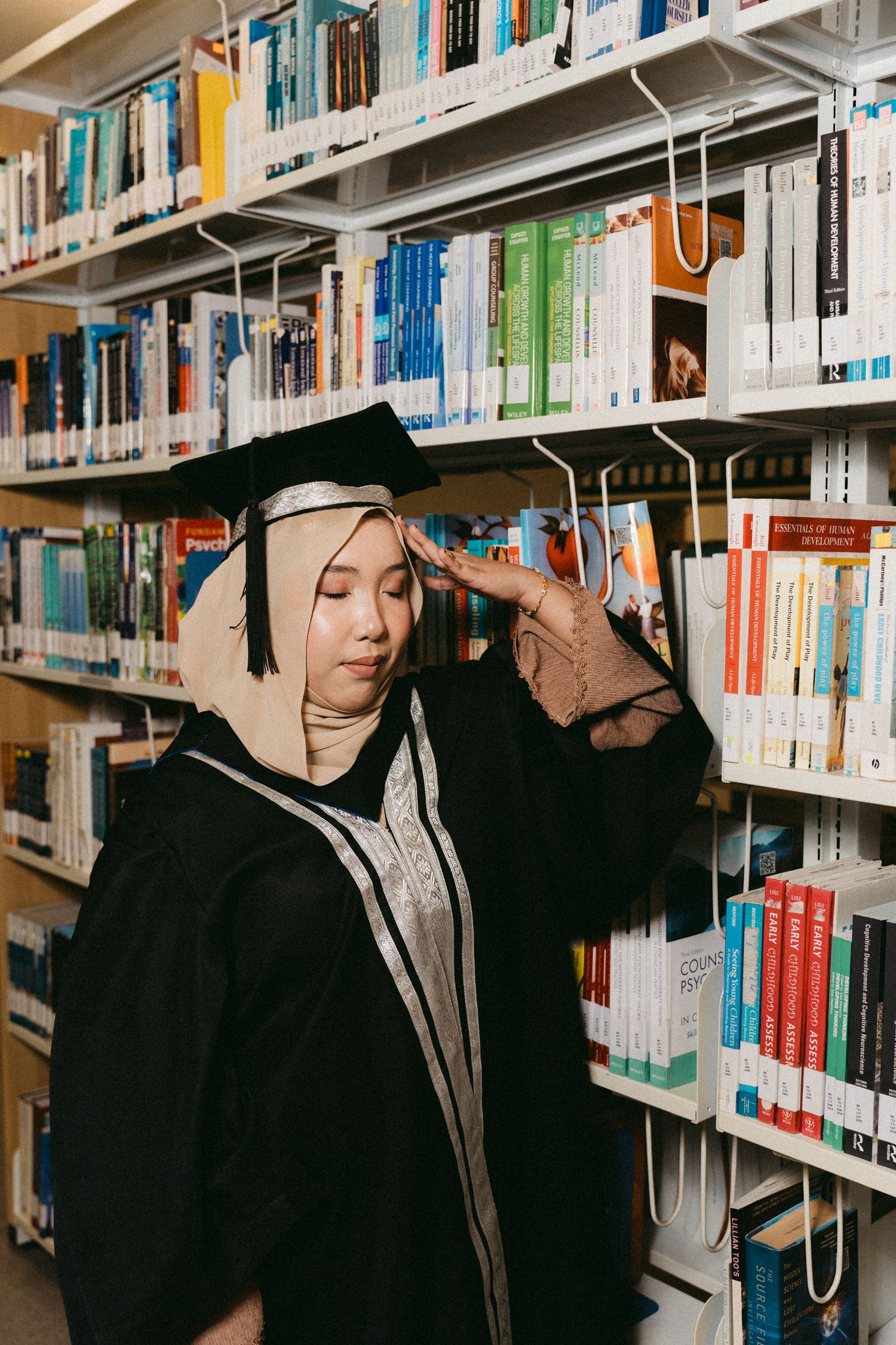 A student wearing a graduation cap and black hoodie stands thoughtfully in front of library bookshelves.