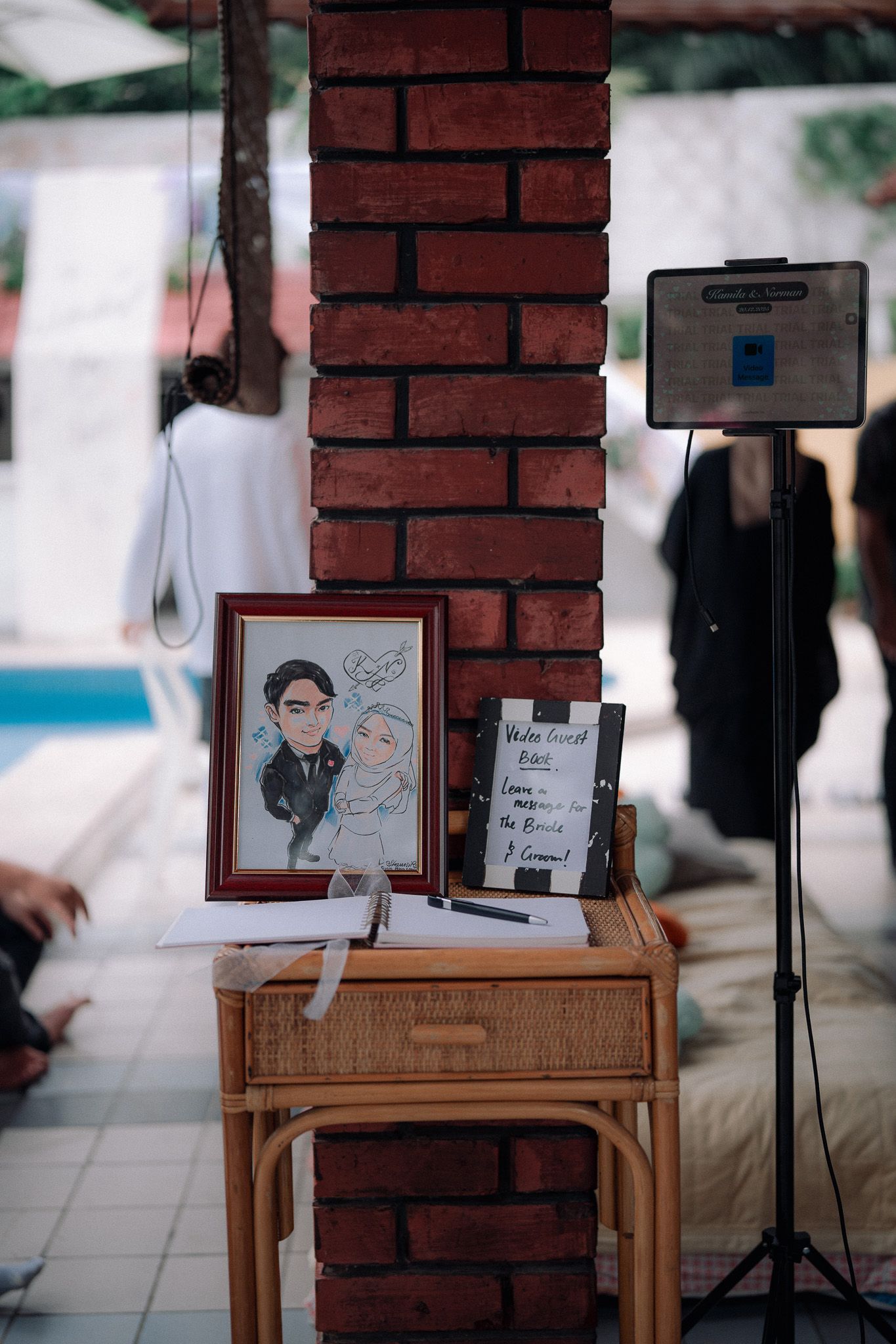A framed caricature of a couple displayed on a rattan table beside a welcome sign at an outdoor wedding venue.