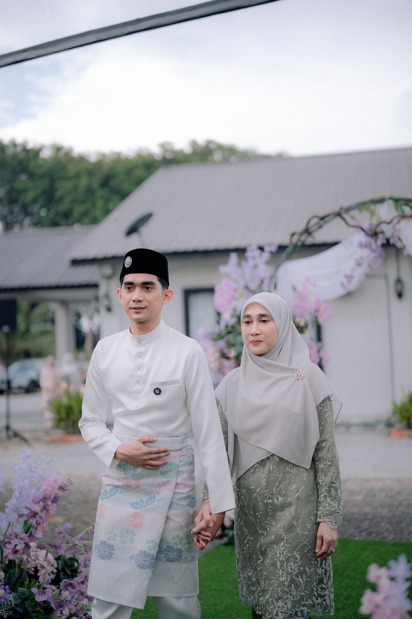 A couple in traditional Malay attire, the man in white baju melayu and the woman in a grey hijab, pose at an outdoor wedding.