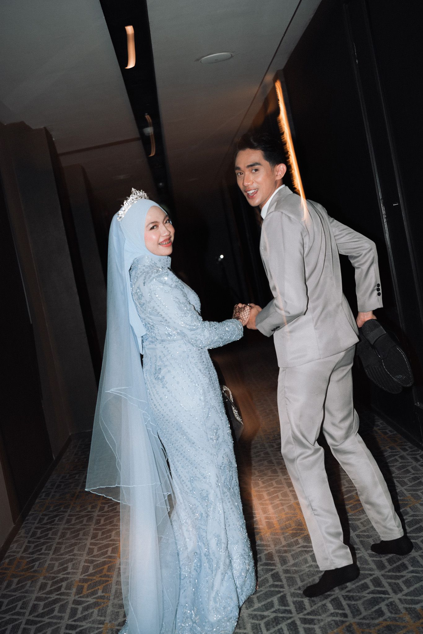 Smiling newlywed Muslim couple in light blue wedding attire hold hands in a hotel corridor.