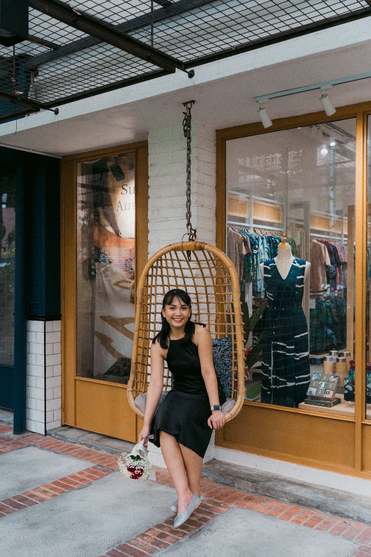 Woman in black dress sitting in suspended rattan chair outside retail store window.