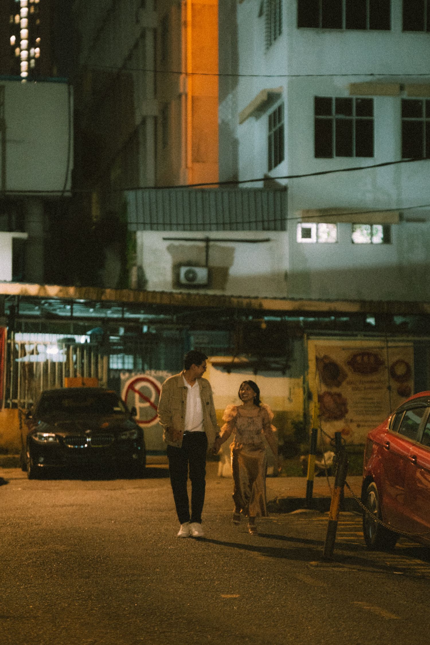 Couple walking through dimly lit urban street at night with parked cars nearby.