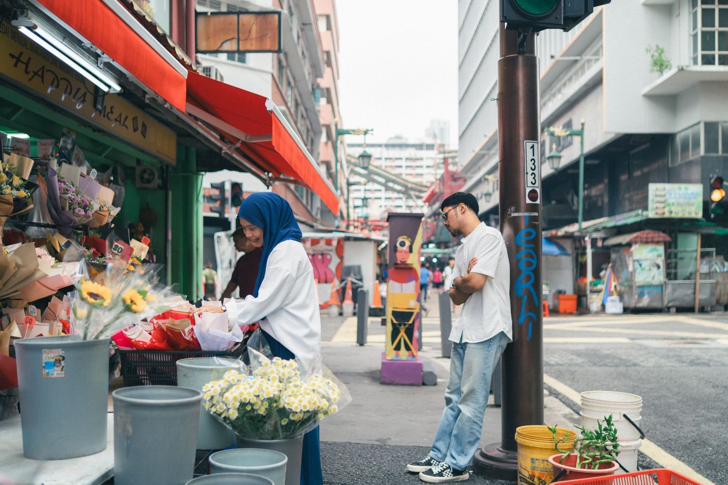 Street vendors and shoppers interact at an urban marketplace with red awnings and fresh produce displays.