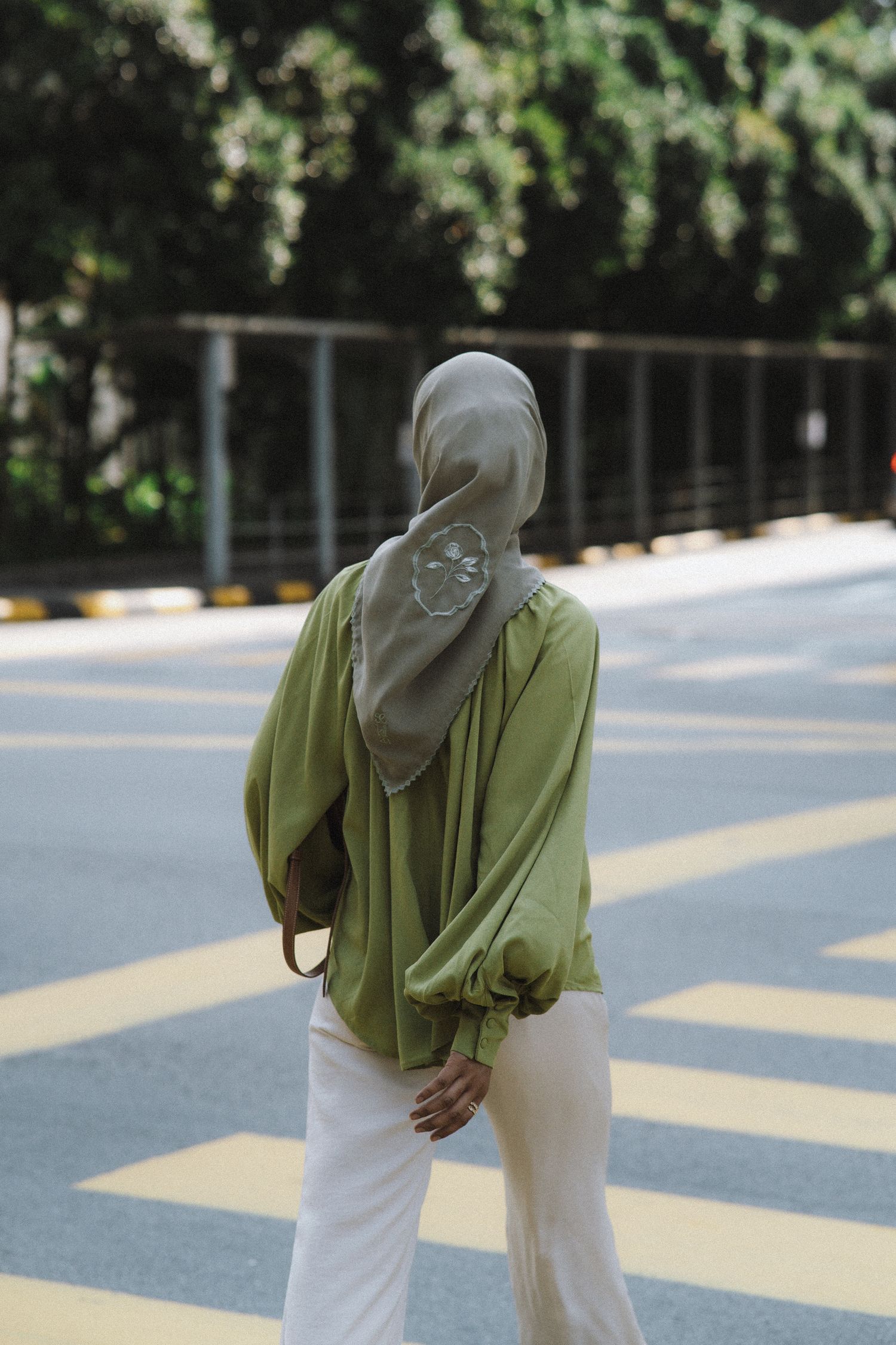 Person in sage green blouse and white pants walks on striped pedestrian crossing.