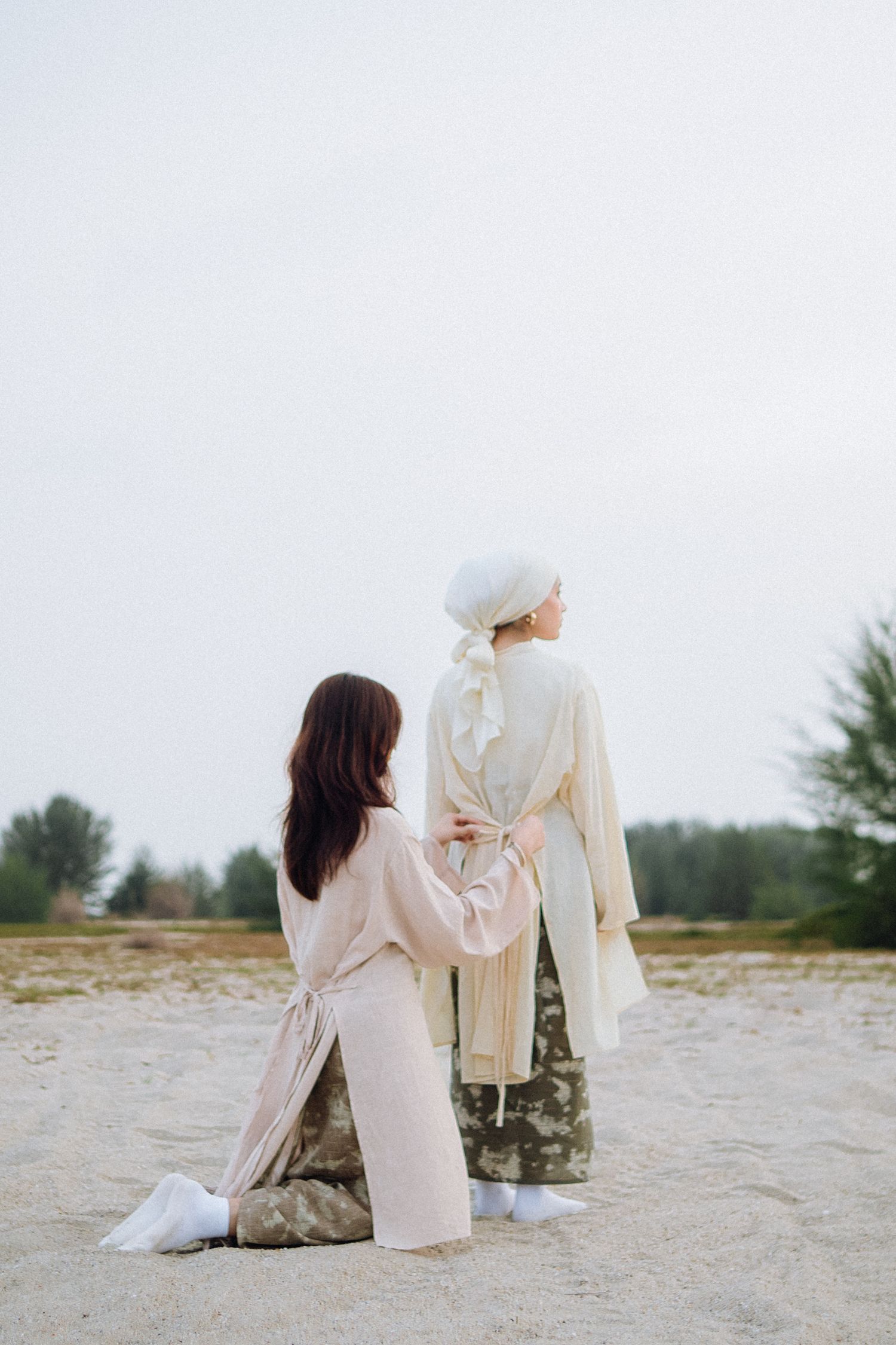 Two people in light-colored clothing sitting and standing on sandy ground against an overcast sky.