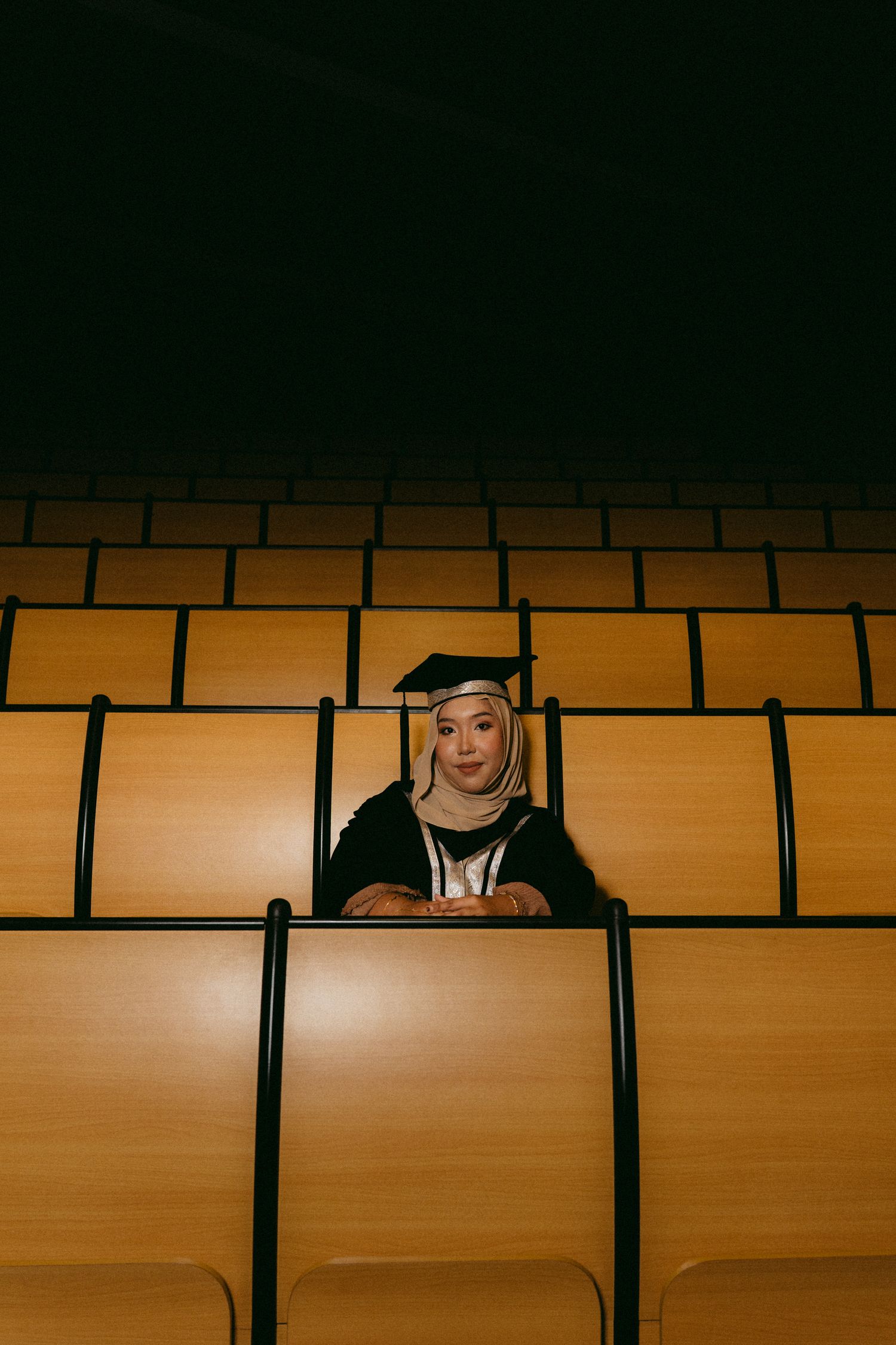 A graduate in academic regalia sits alone in an empty classroom lecture hall with orange lighting.