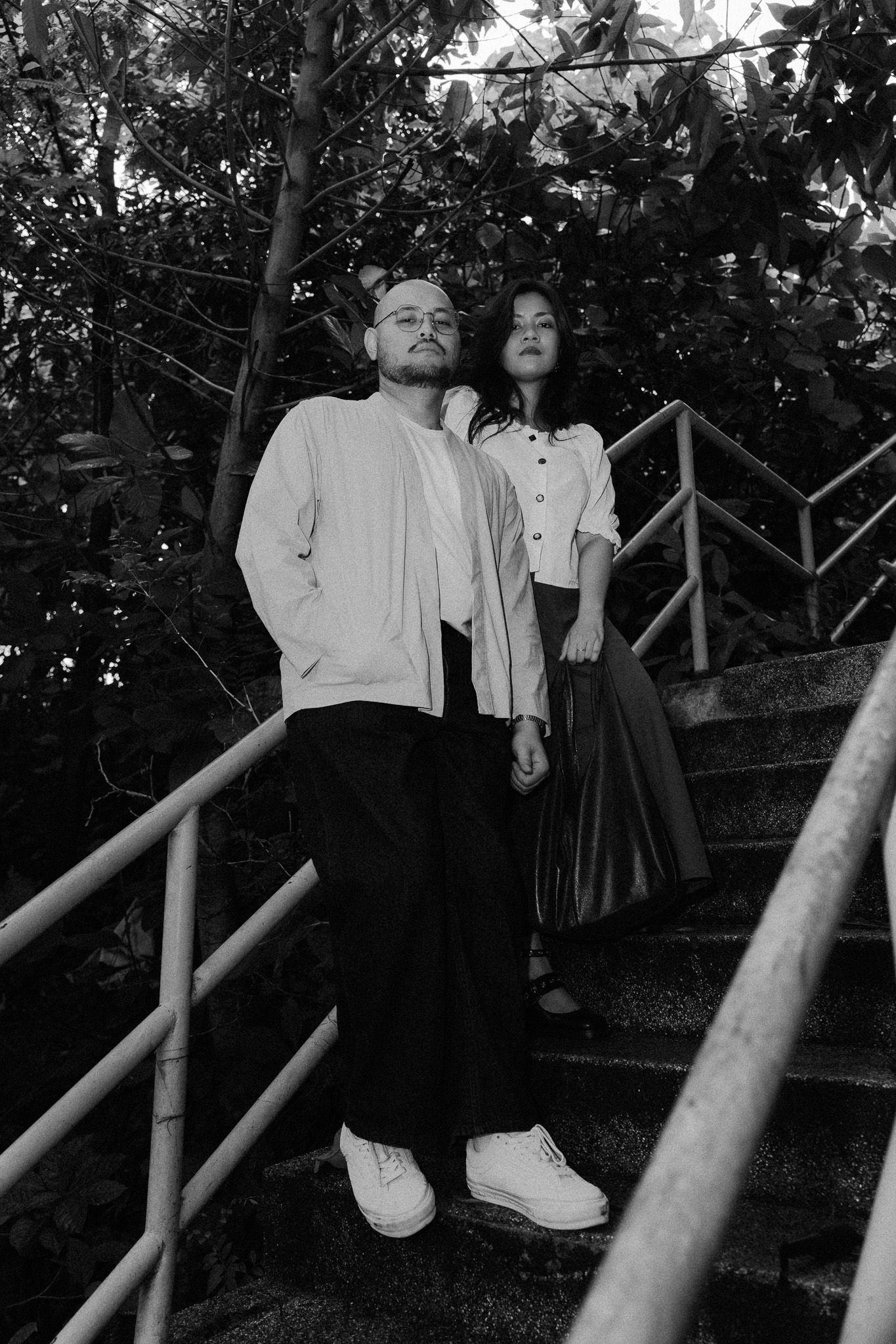 Black and white photo of a stylish couple posing on outdoor stairs surrounded by lush green trees.
