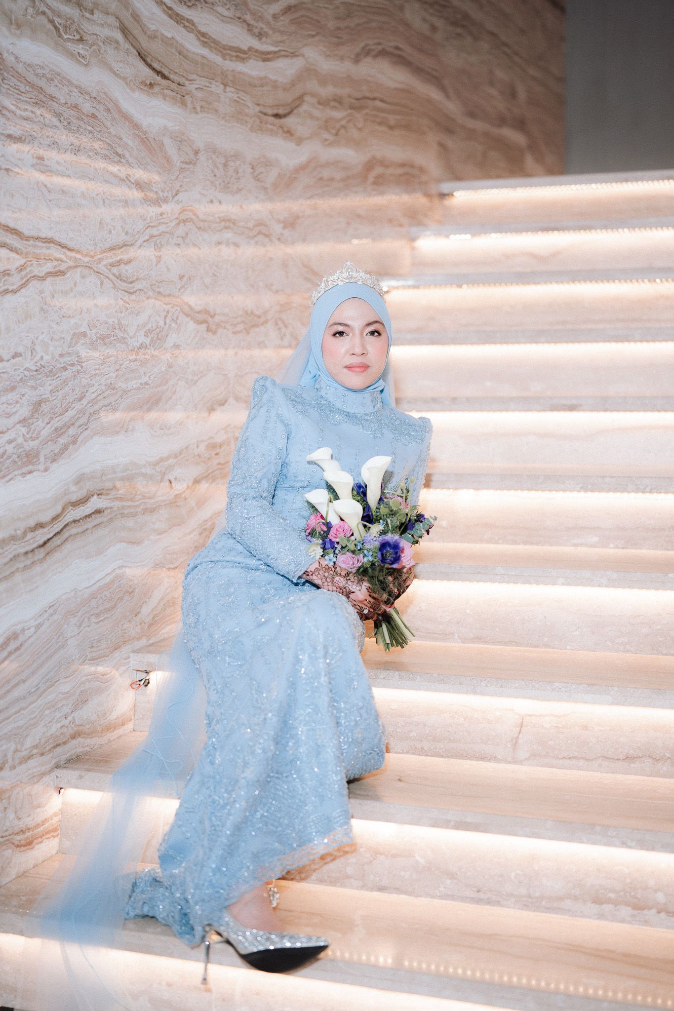 A bride in a light blue hijab and sequined gown sits on illuminated marble stairs holding a floral bouquet.