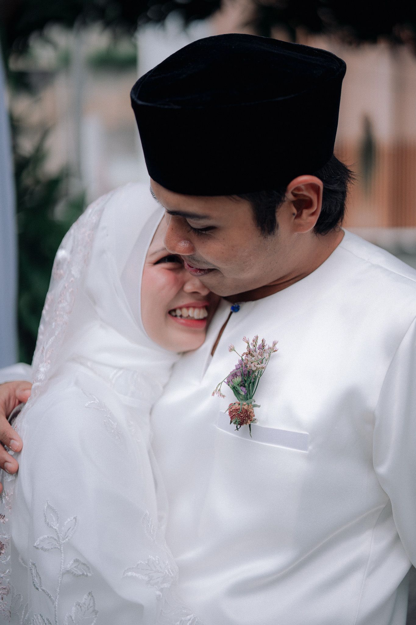 Muslim couple in white wedding attire embracing, groom wearing black songkok hat with boutonniere, bride in white hijab smiling.