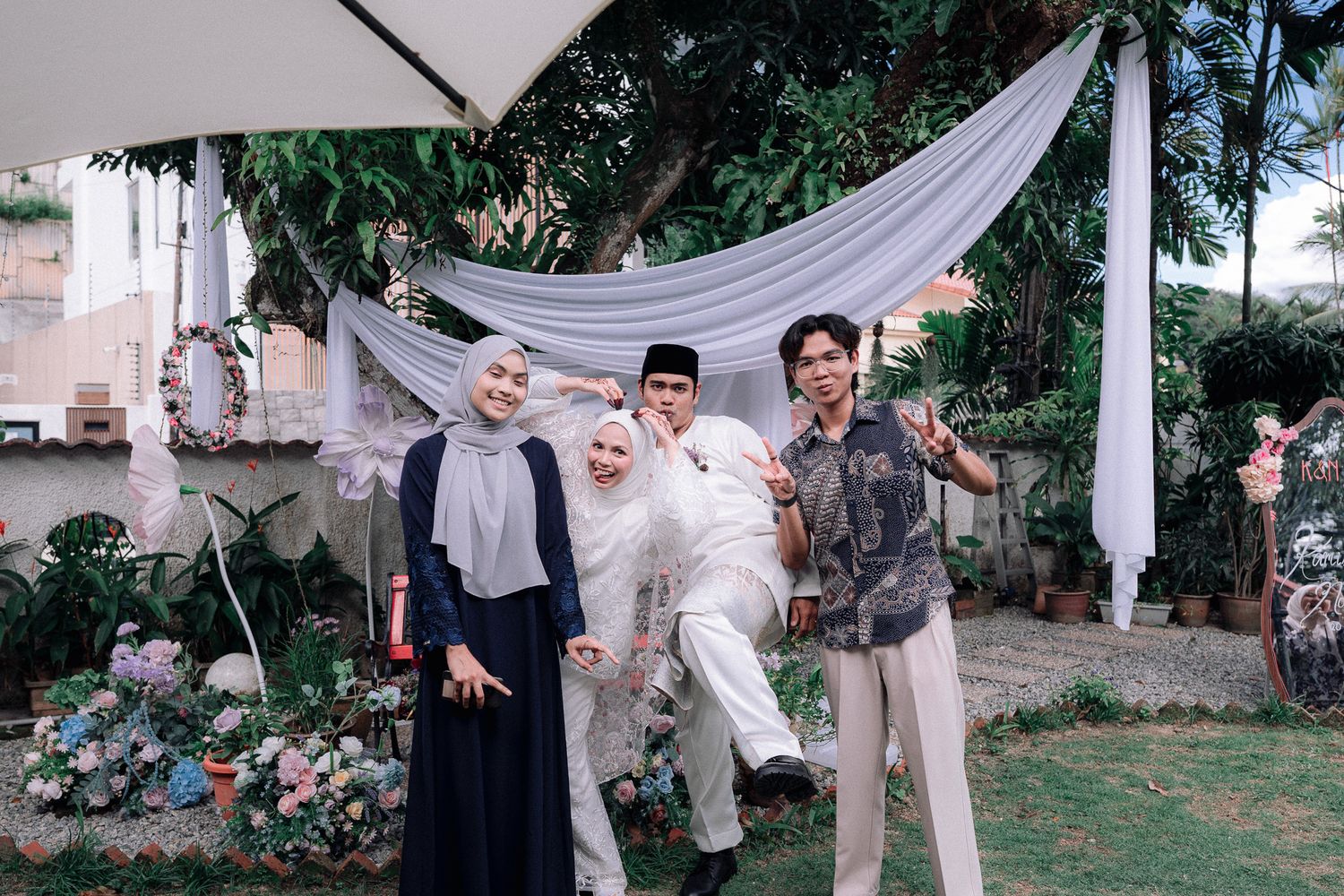 Group of four friends posing together outdoors at a garden celebration with white draping and lush greenery.