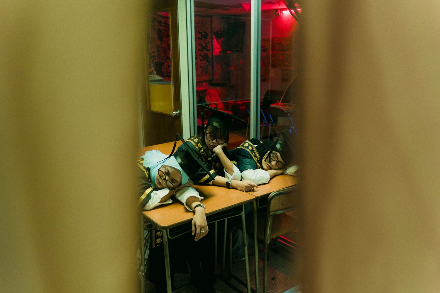 Two exhausted students sleeping at a desk in a dimly lit room with red ambient lighting.