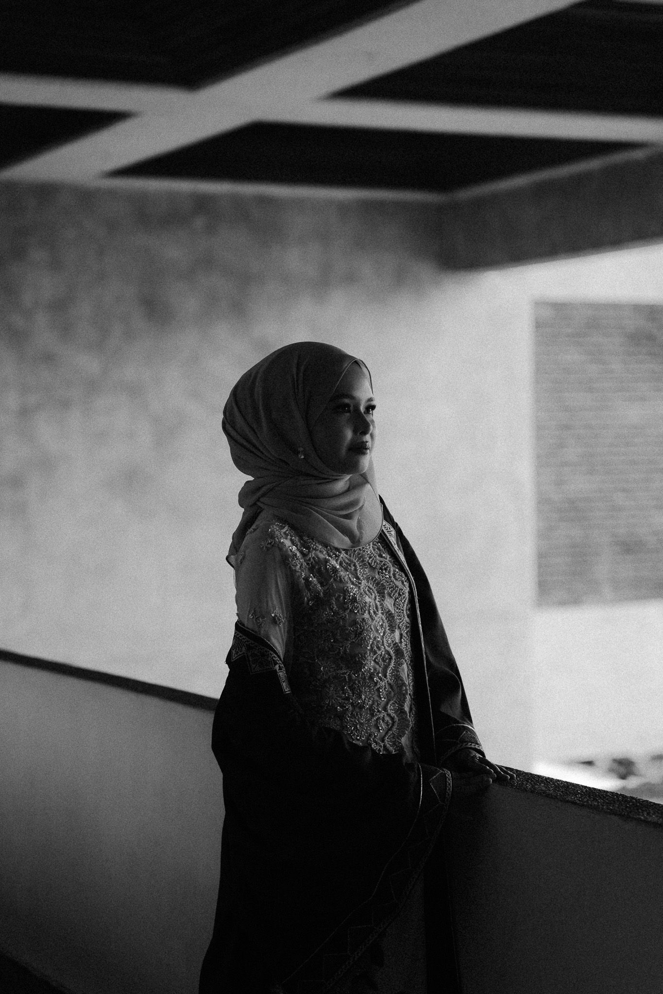 Black and white portrait of a woman wearing a hijab and lace top, standing gracefully in a dimly lit architectural space.