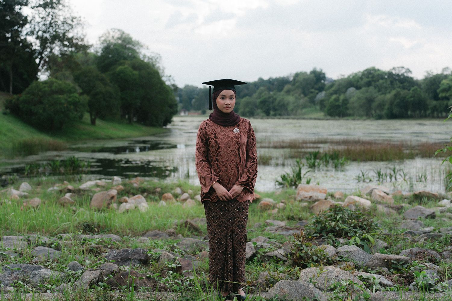 A young woman in batik dress and graduation cap stands proudly by a serene river surrounded by lush greenery.