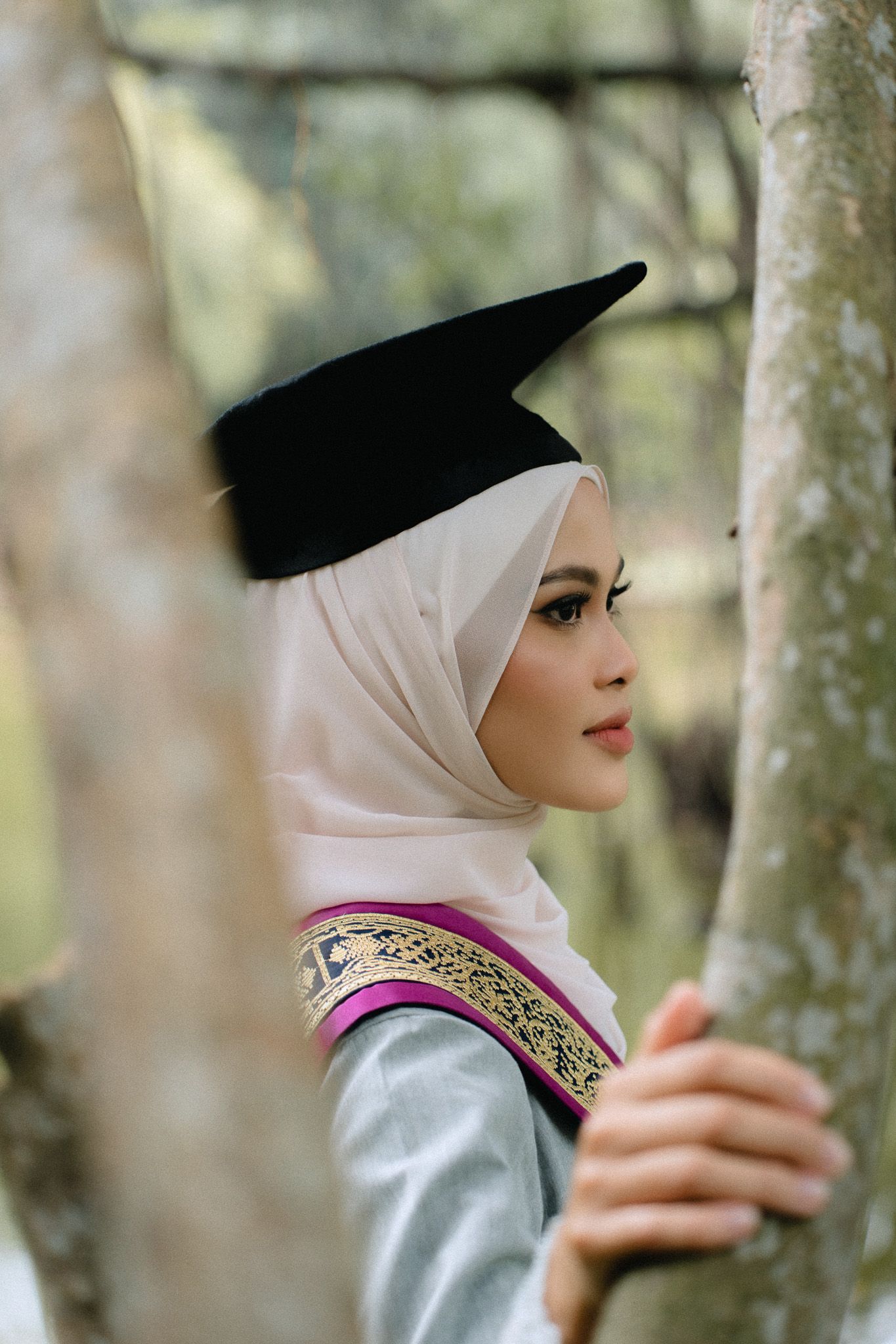 Young Muslim woman in hijab and graduation cap poses thoughtfully among trees in traditional attire.