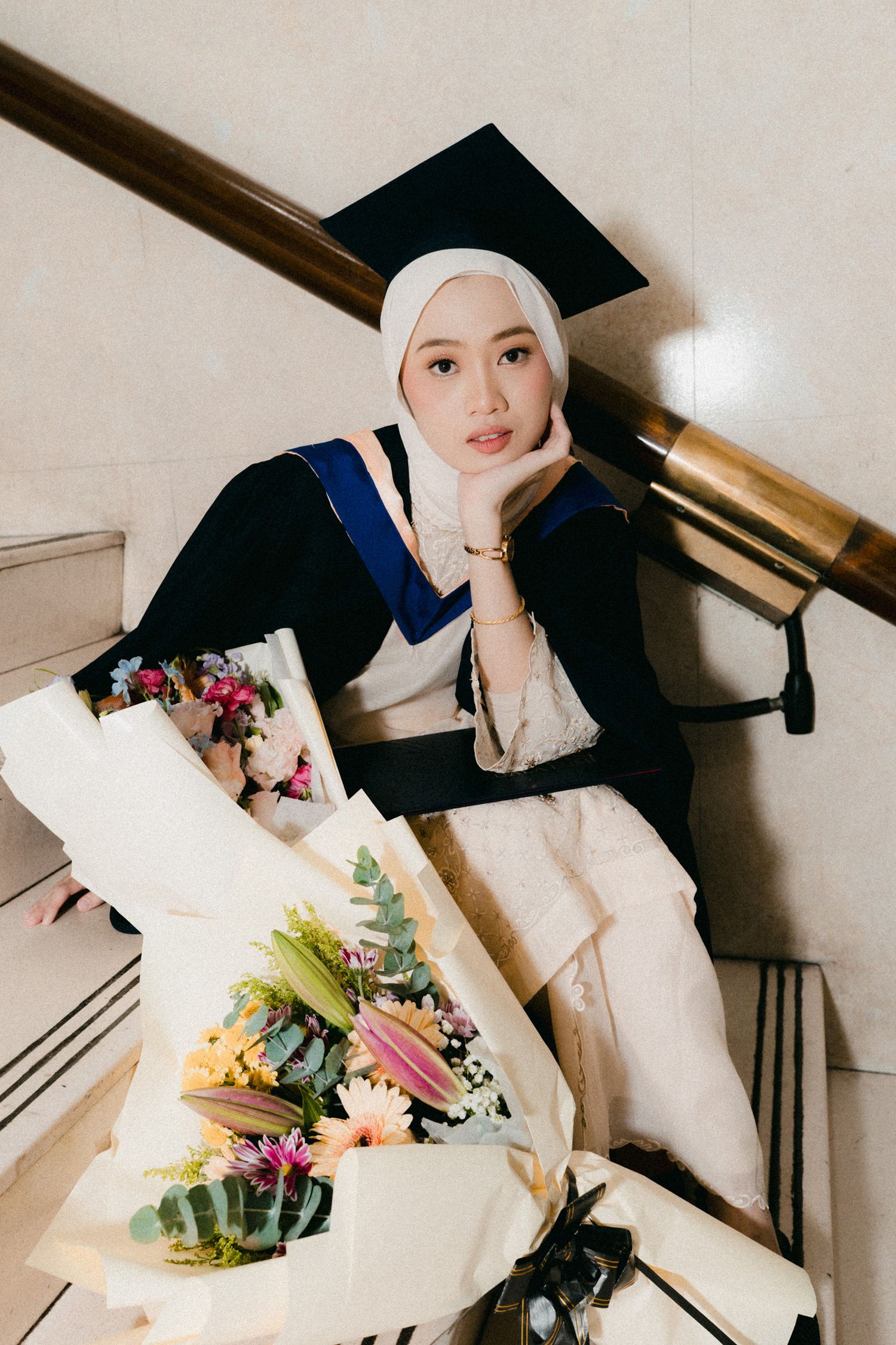Graduate wearing black cap and white hijab holds colorful floral bouquets while posing for graduation photos.