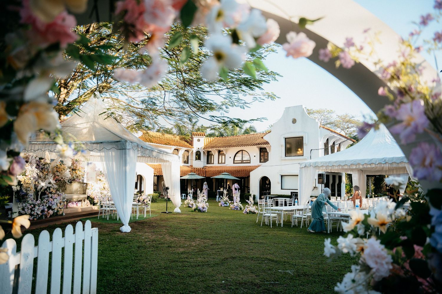 A white outdoor wedding tent setup on a green lawn surrounded by trees and flowers during sunset.