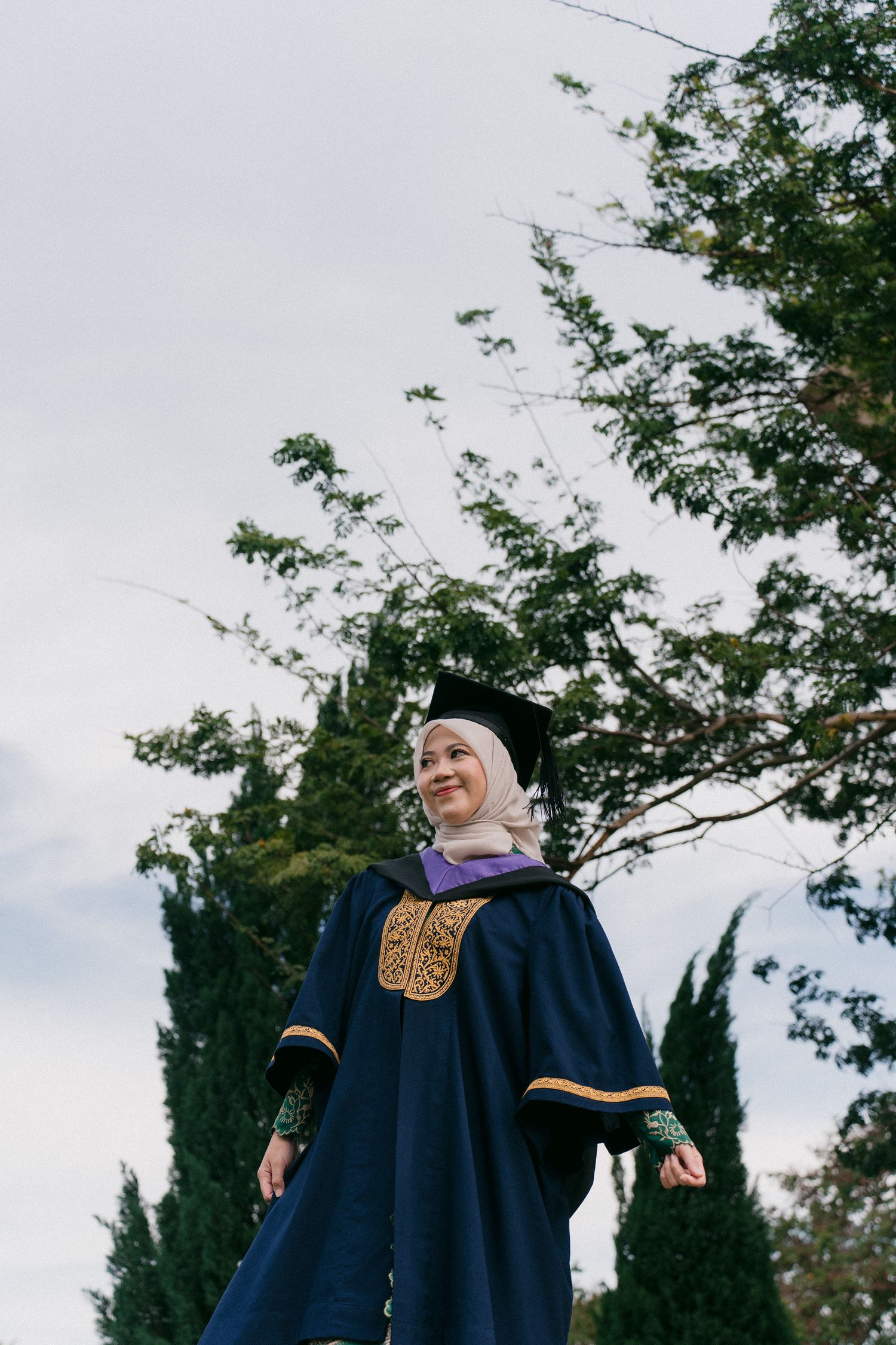 Graduates in navy blue academic regalia stand together outdoors under green trees during commencement ceremony.