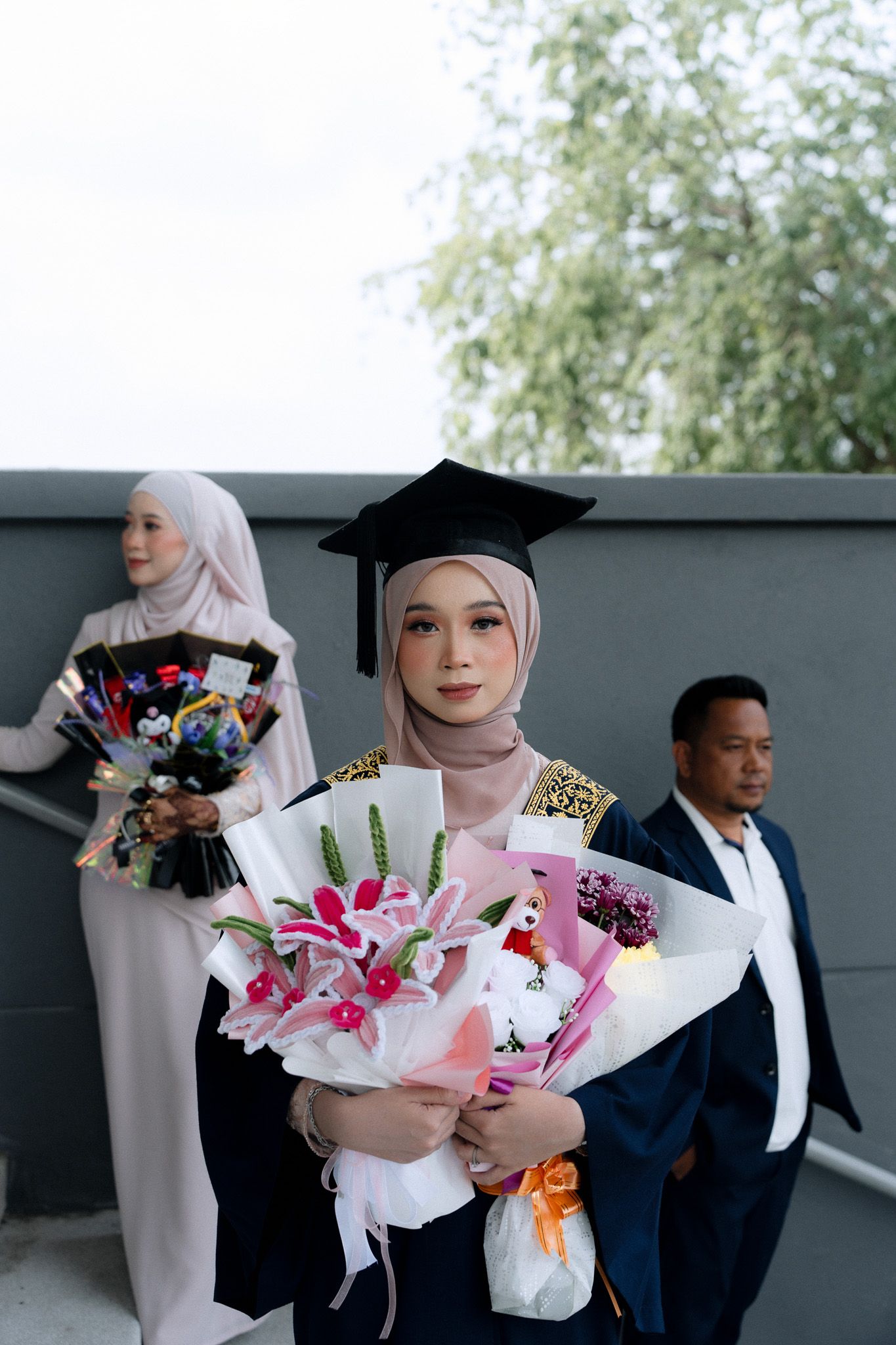Muslim graduate in cap and gown holds flower bouquet, with family members standing behind her at graduation ceremony.