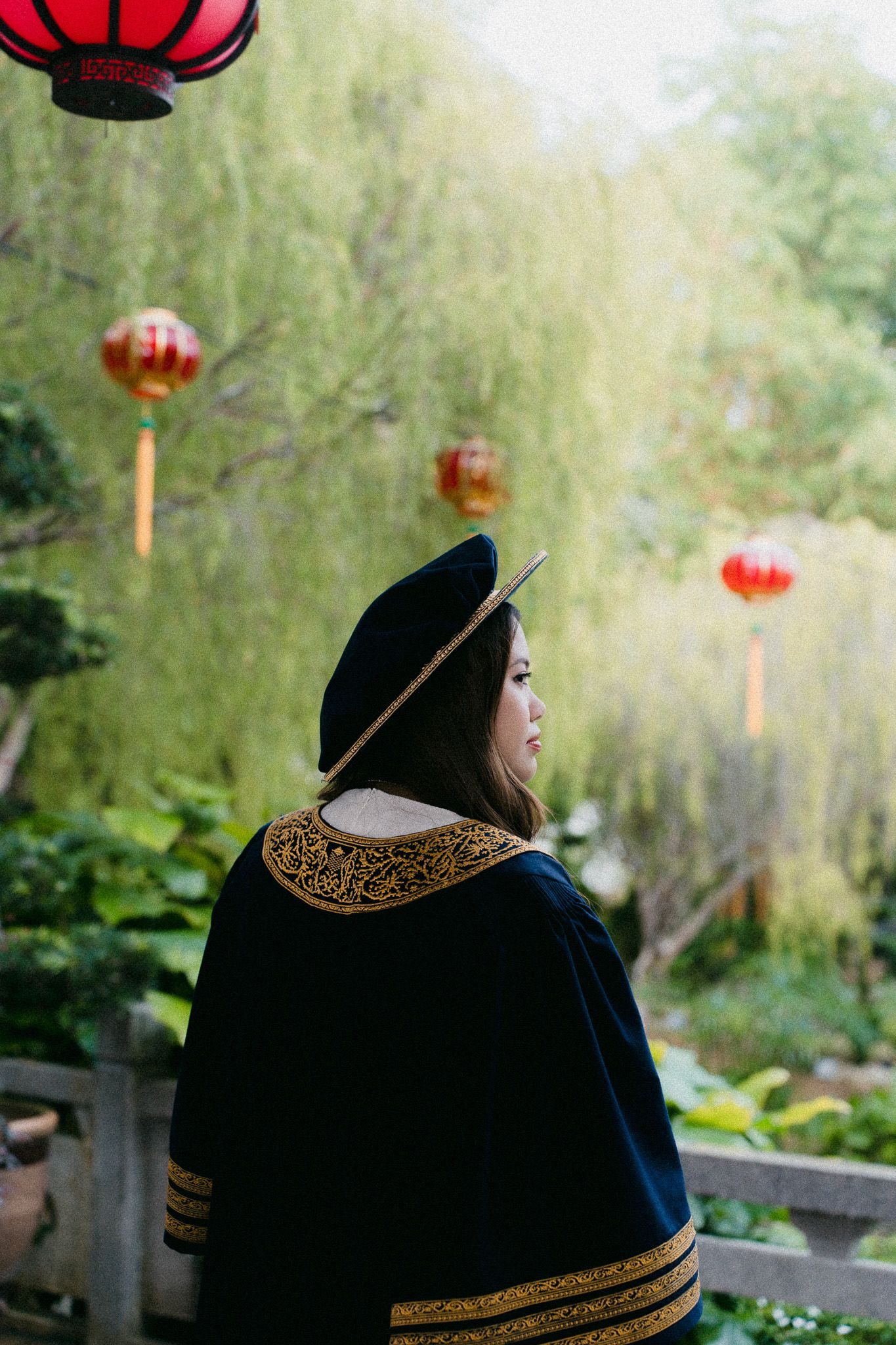 Woman in graduation gown and cap stands in a Chinese garden with red lanterns hanging from trees.