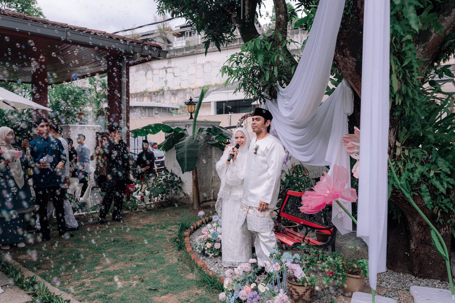 A couple in white wedding attire stands outdoors amid falling confetti, surrounded by guests and floral decorations.
