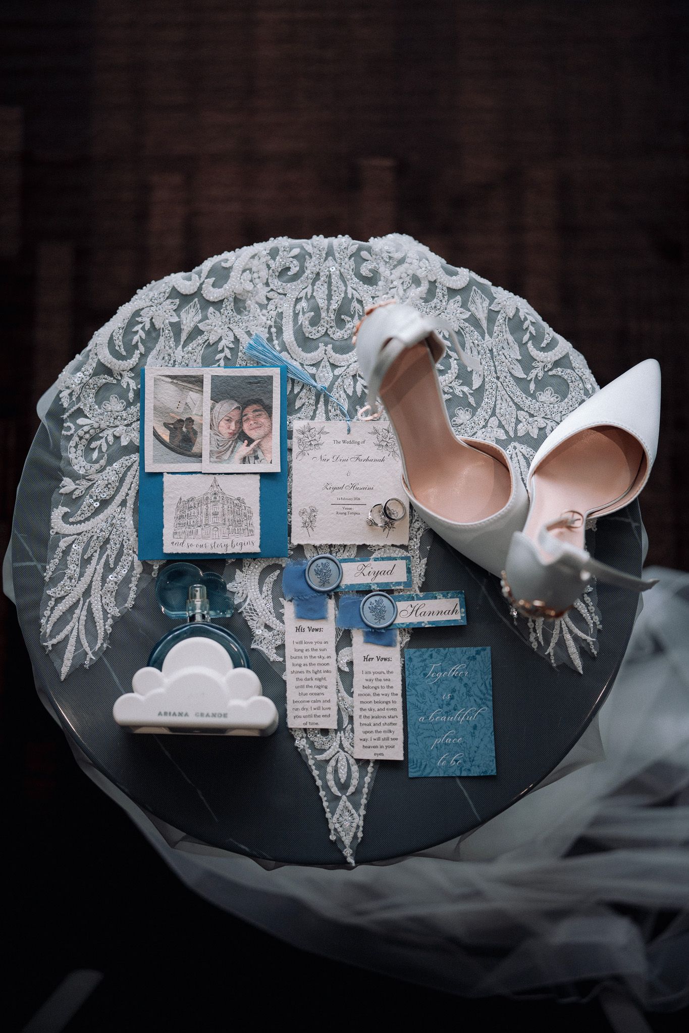 Elegant wedding flat lay with white heels, blue stationery, photos, and lace veil on a dark round table.