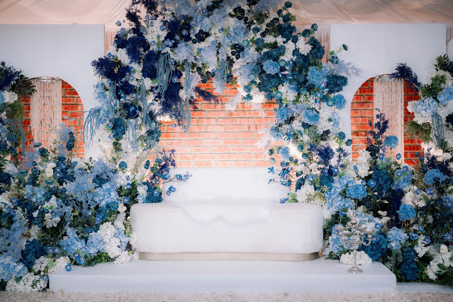 Elegant wedding backdrop with blue and white floral arrangements framing a white sofa against a brick wall.