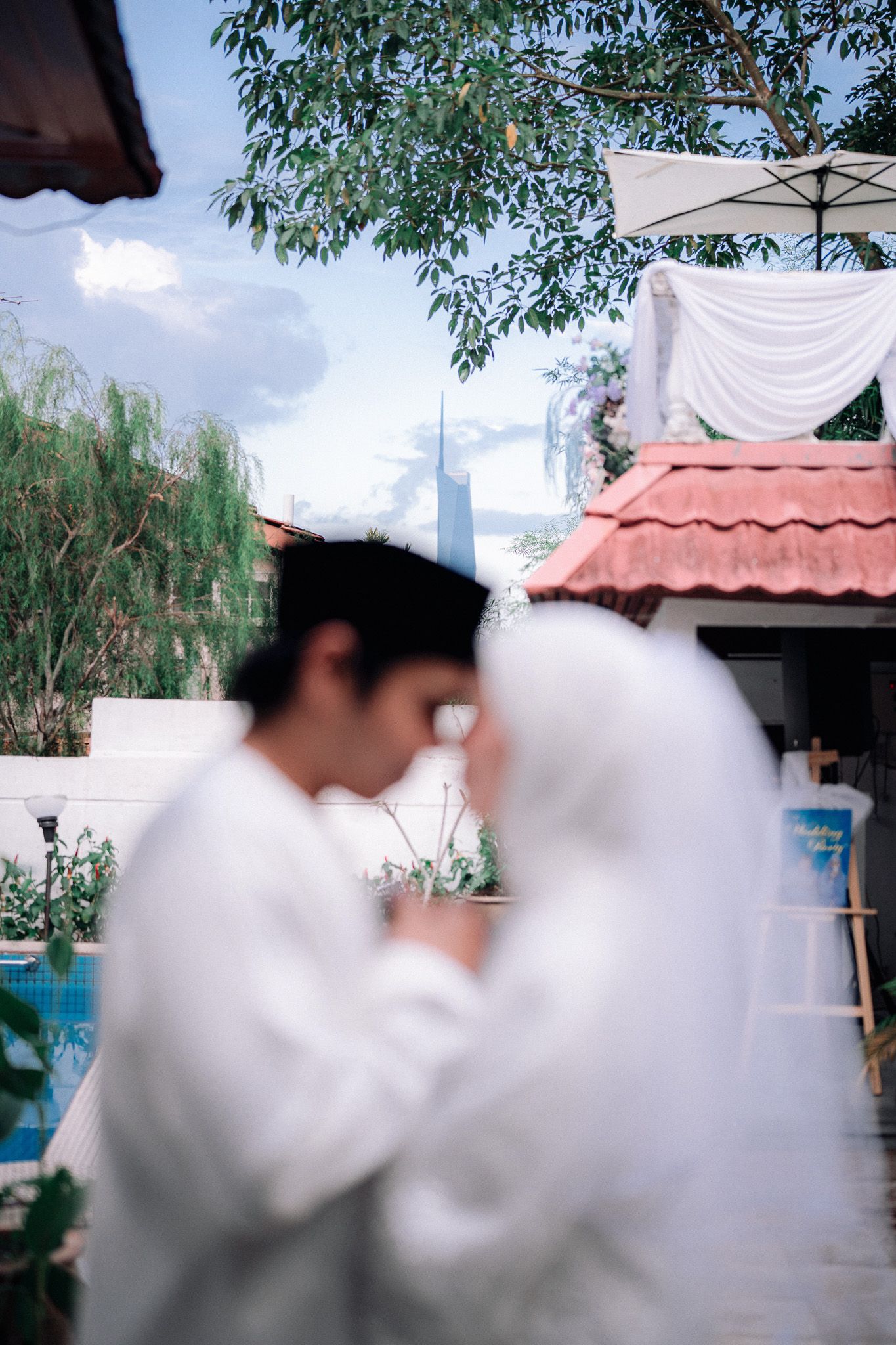 Muslim couple in white wedding attire sharing an intimate moment outdoors near a pool with lush greenery.