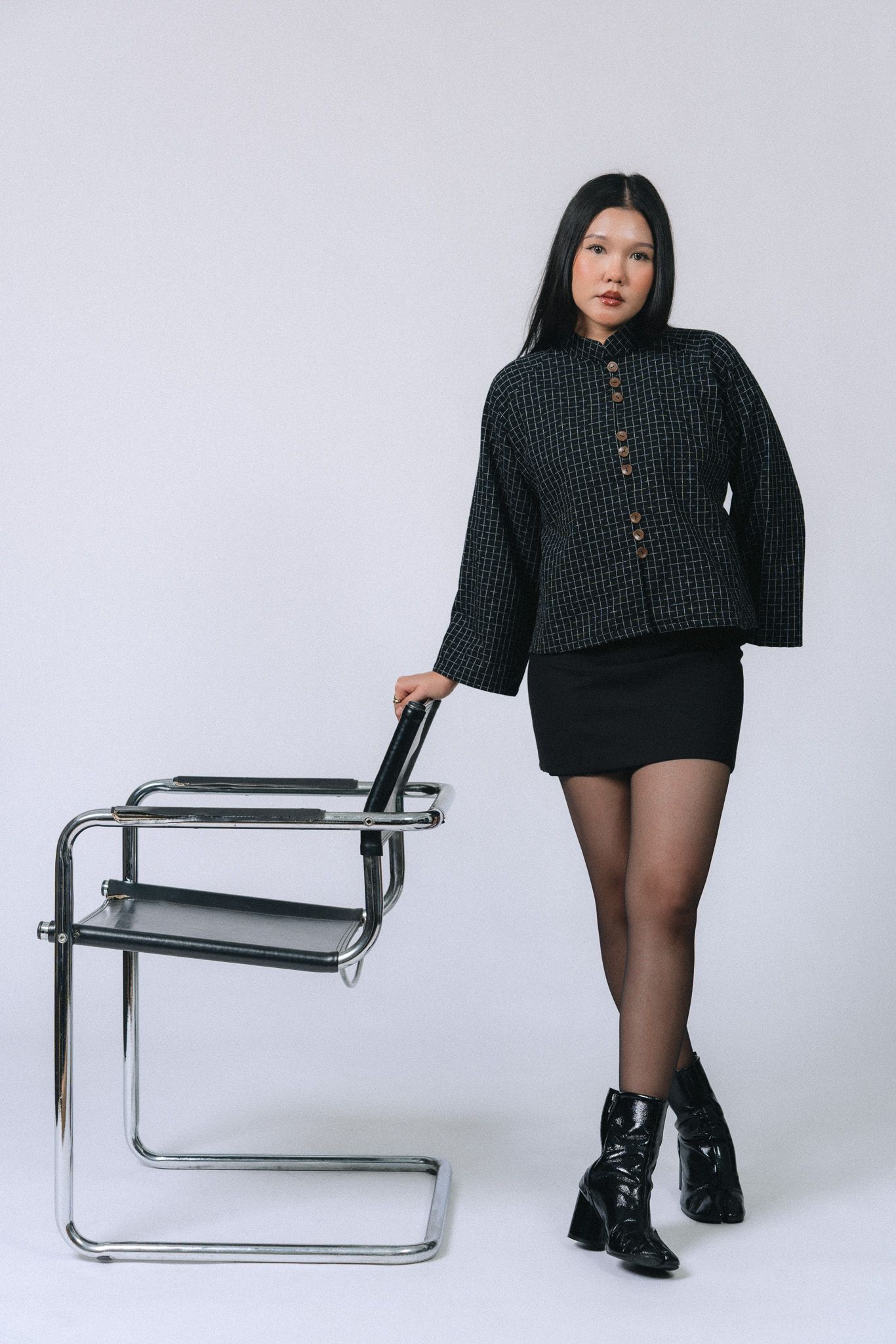 Young woman in black mini skirt, checkered jacket, and patent boots poses beside a modern chrome cantilever chair.