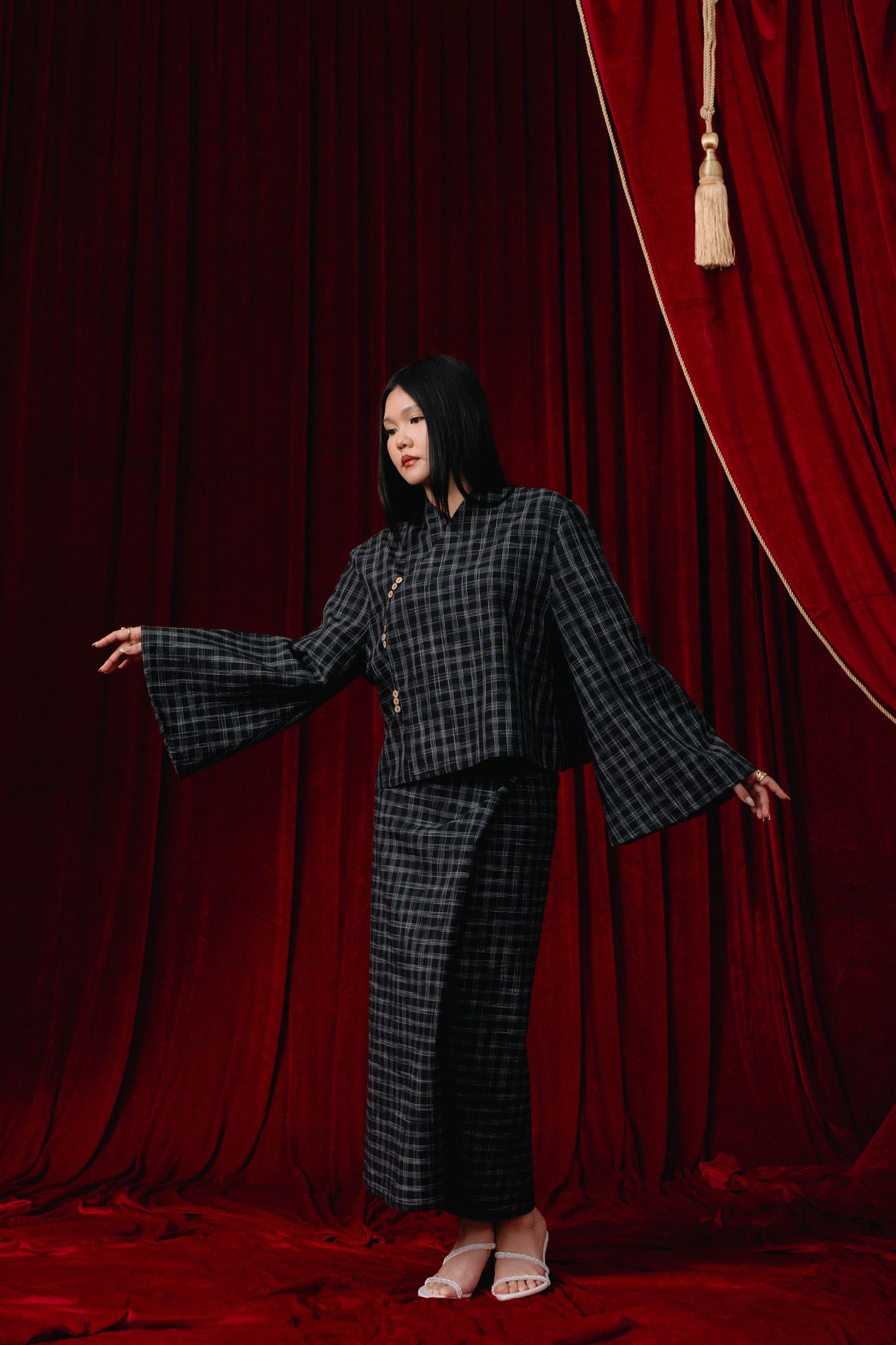 Woman in plaid Chinese-style suit with flared sleeves poses against rich red velvet curtain backdrop.