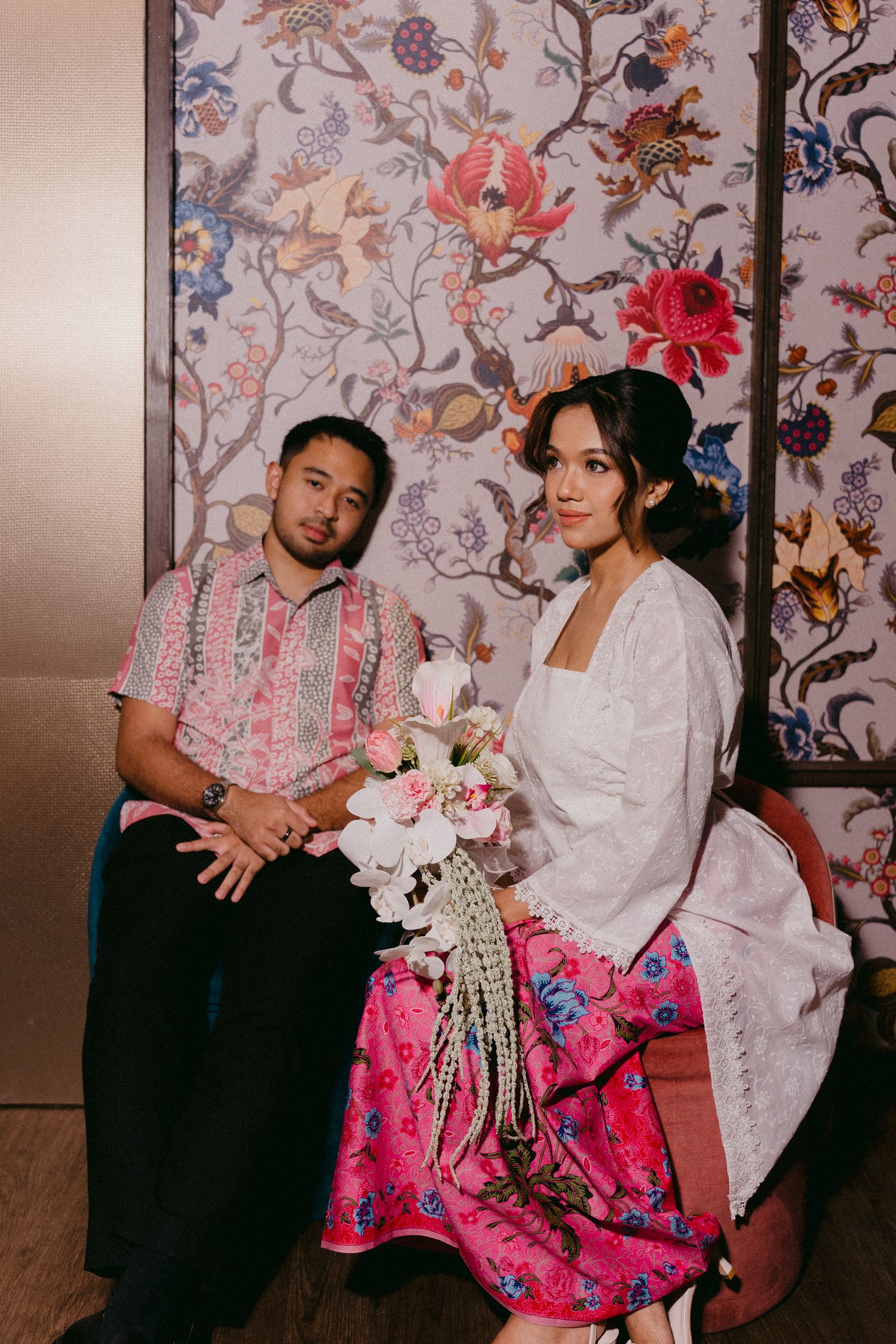 Couple in traditional Asian-inspired attire poses against floral wallpaper with pink and white flower bouquet.