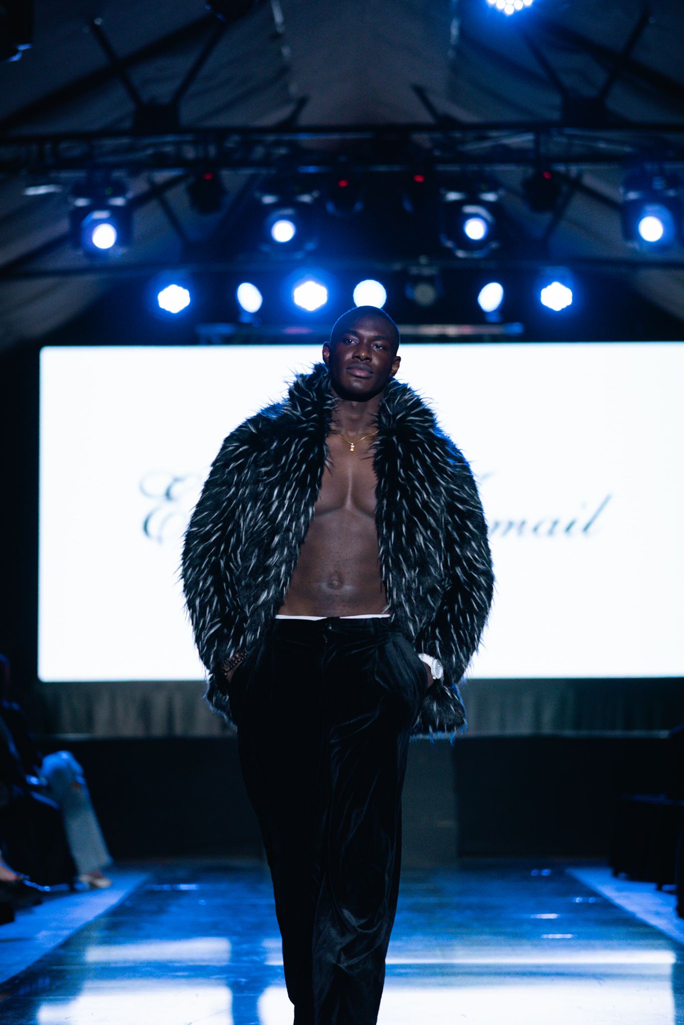 Model wearing dramatic black feathered coat walks runway under blue stage lights.