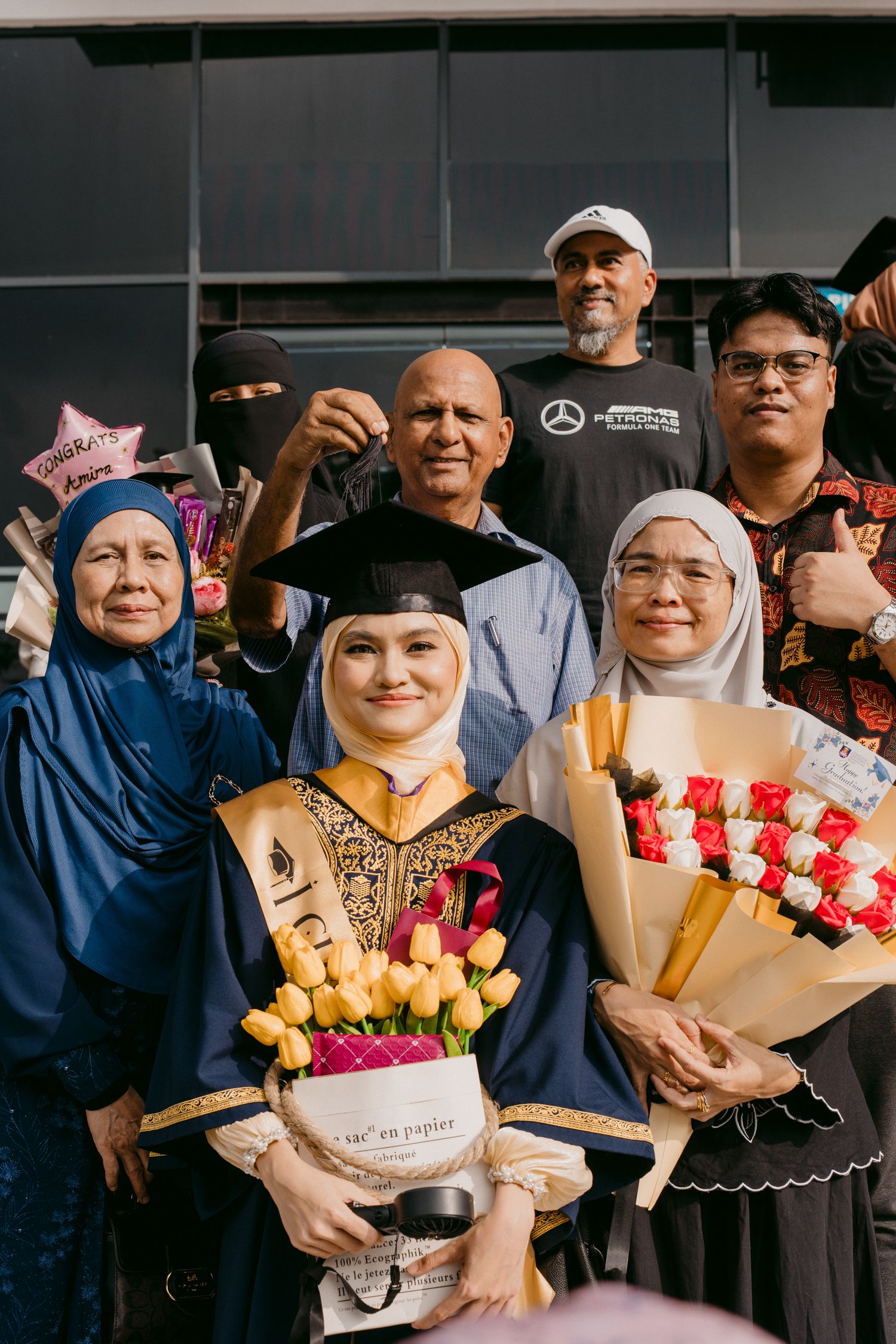 A graduation celebration scene with a group wearing traditional clothing and holding bouquets of flowers and celebration props.