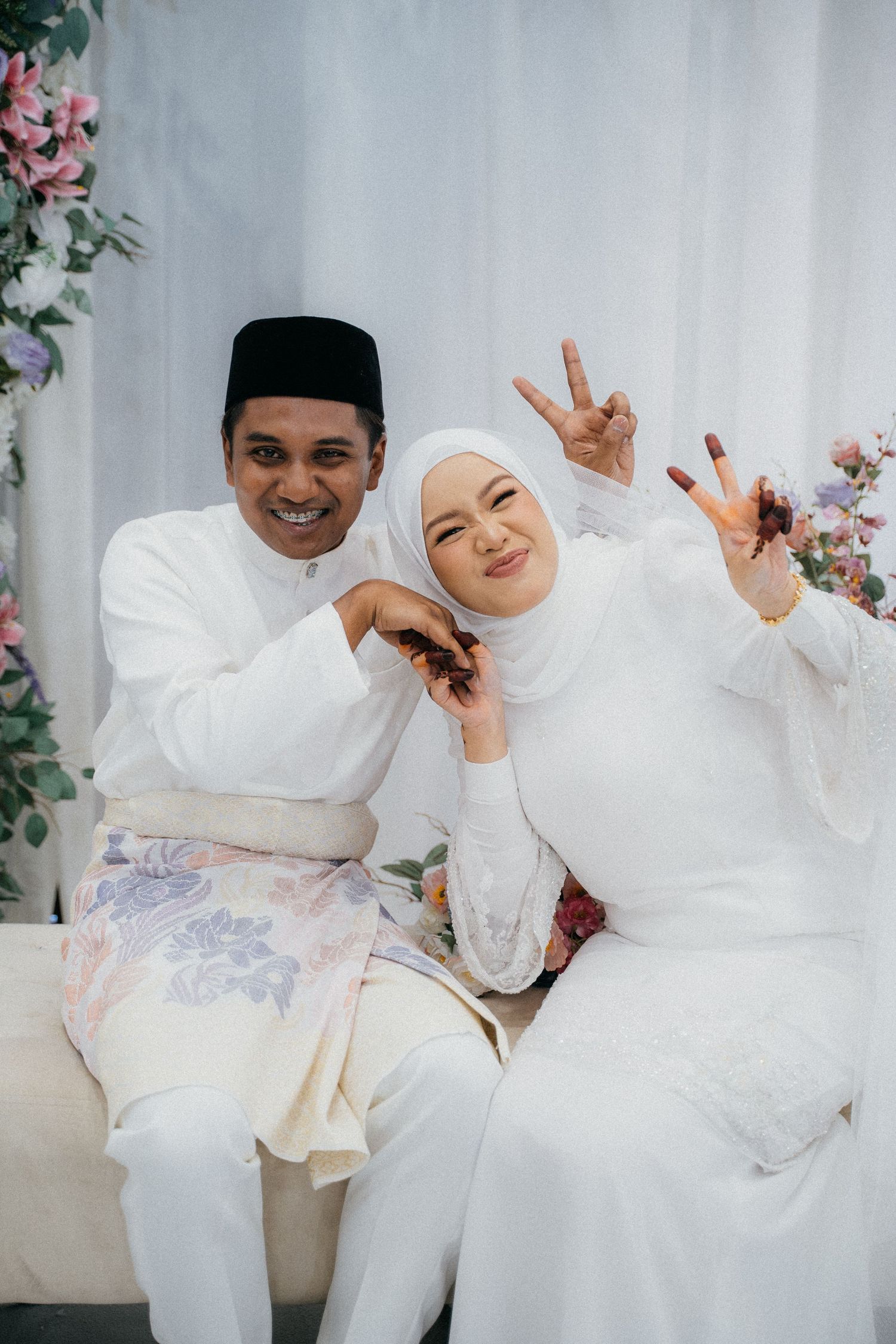 A joyful couple in traditional Malay wedding attire pose together making peace signs against a floral backdrop.