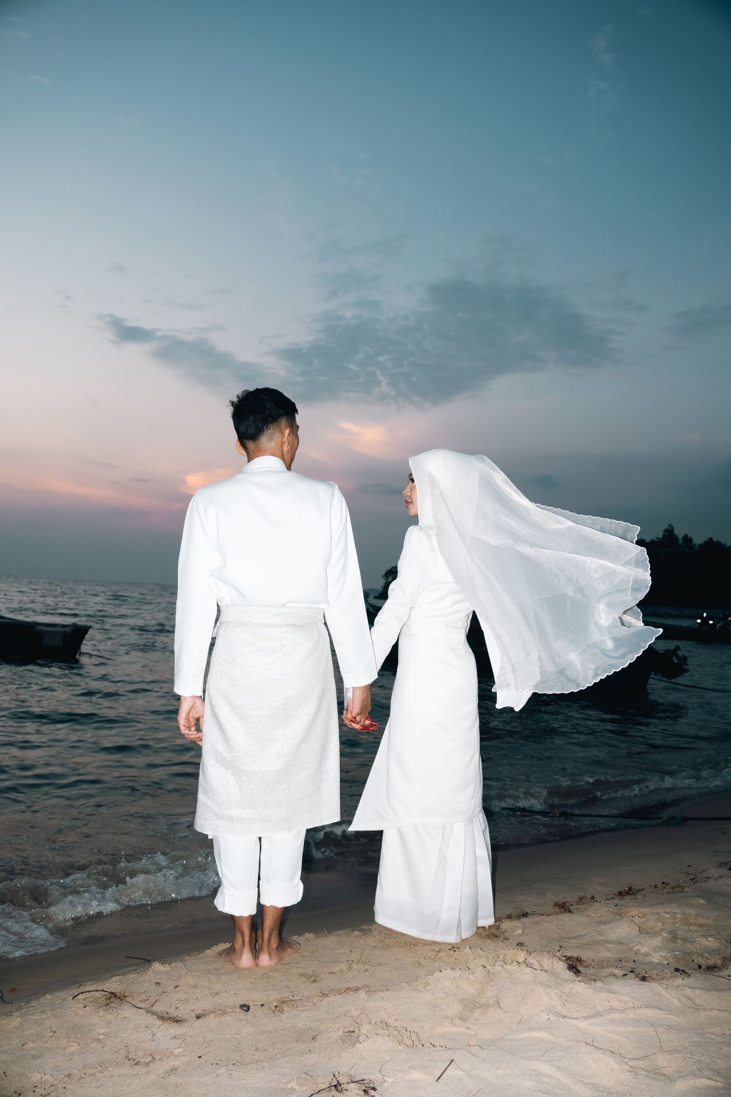 A couple in white attire holding hands on a beach at sunset with flowing veil caught in the ocean breeze.