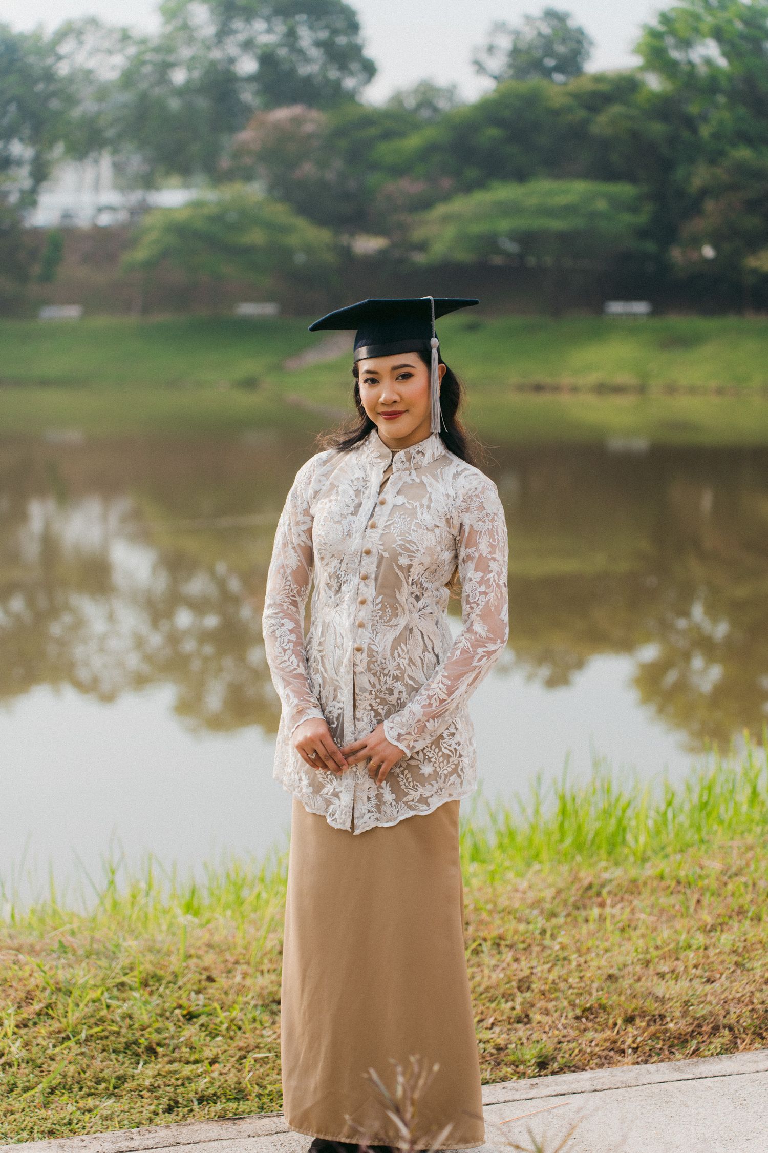 A graduate in a cap and gown poses by a lake wearing a beige skirt and white lace top for graduation photos.