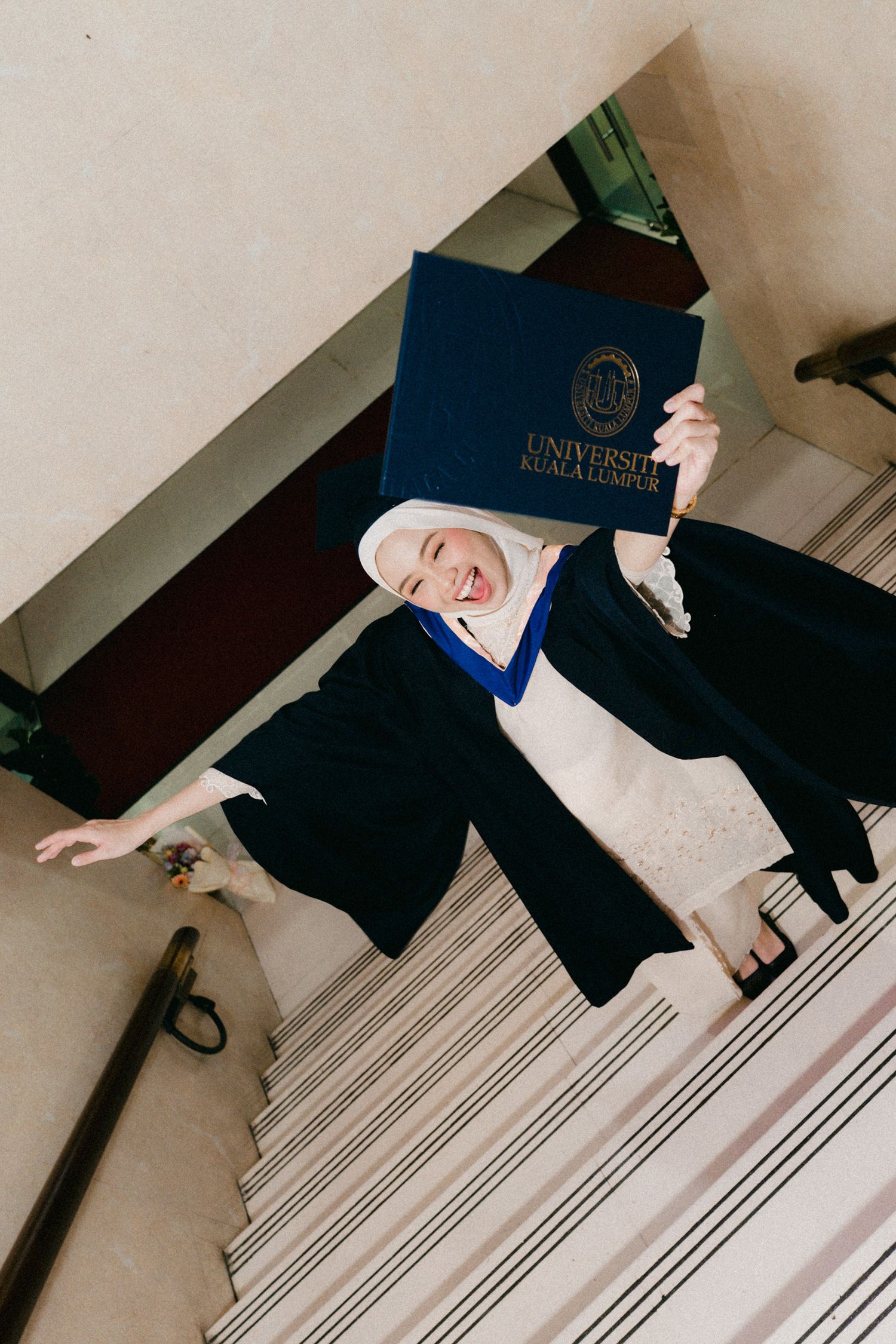 Graduate in academic regalia celebrates with diploma while standing on a striped staircase.