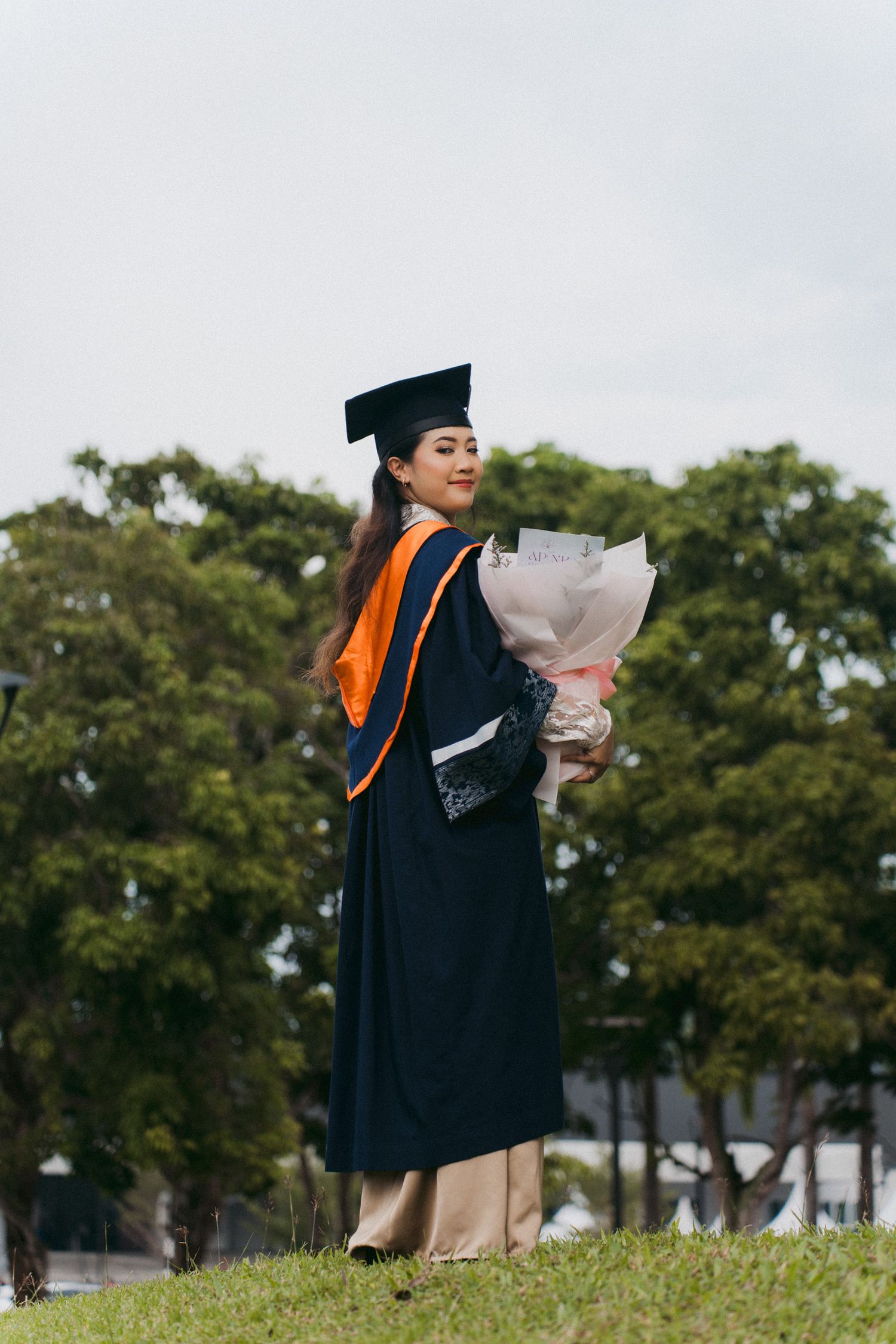 Graduate in black cap and gown with orange stole poses on grassy hill during graduation photoshoot.